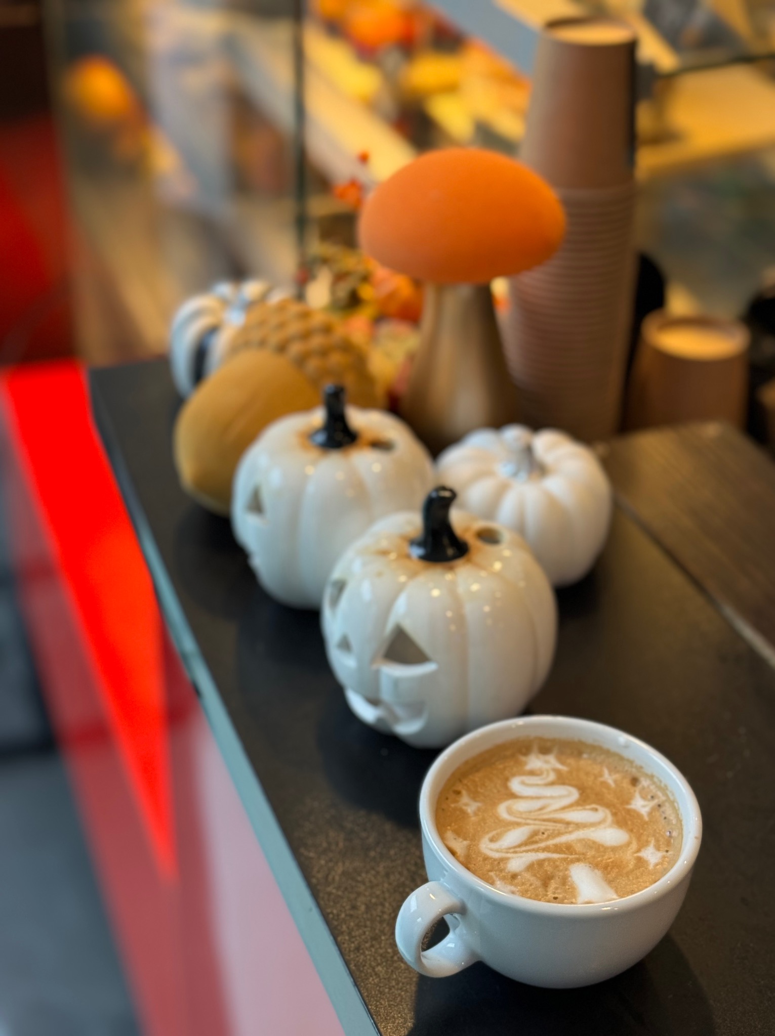 Jesienne dekoracje: białe dynie z wyciętymi twarzami i latte art z motywem choinki w białej filiżance na ciemnym blacie.