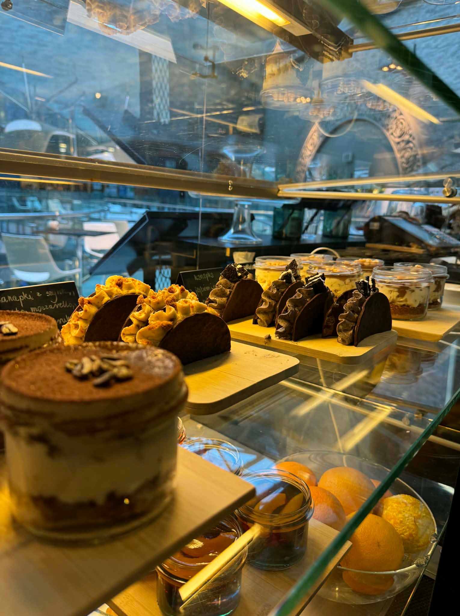 Witryna cukiernicza z deserami: tiramisu, tartaletki z kremem i ciastka w słoiczkach. W tle widoczne odbicie wnętrza kawiarni.