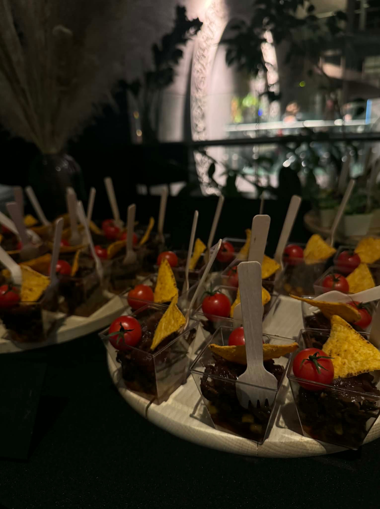 Elegancko podane porcje chili con carne w małych, przezroczystych naczyniach, udekorowane pomidorkami cherry i nachos, na drewnianej tacy. Dekoracje roślinne w tle.