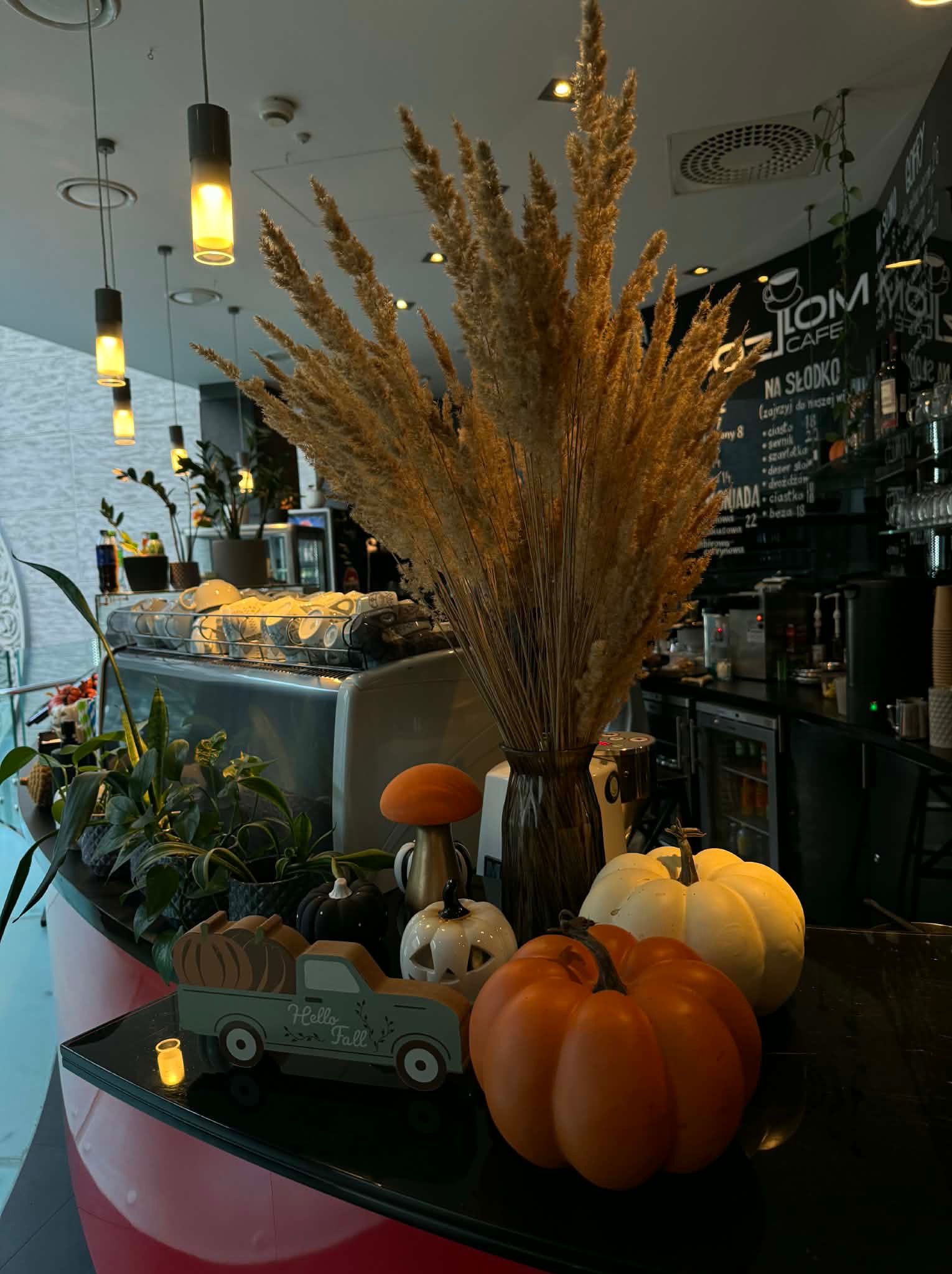 Jesienna dekoracja w kawiarni: dynie, trawa pampasowa w wazonie, drewniany samochodzik z napisem 'Hello Fall', ciepłe oświetlenie, menu na tablicy w tle.
