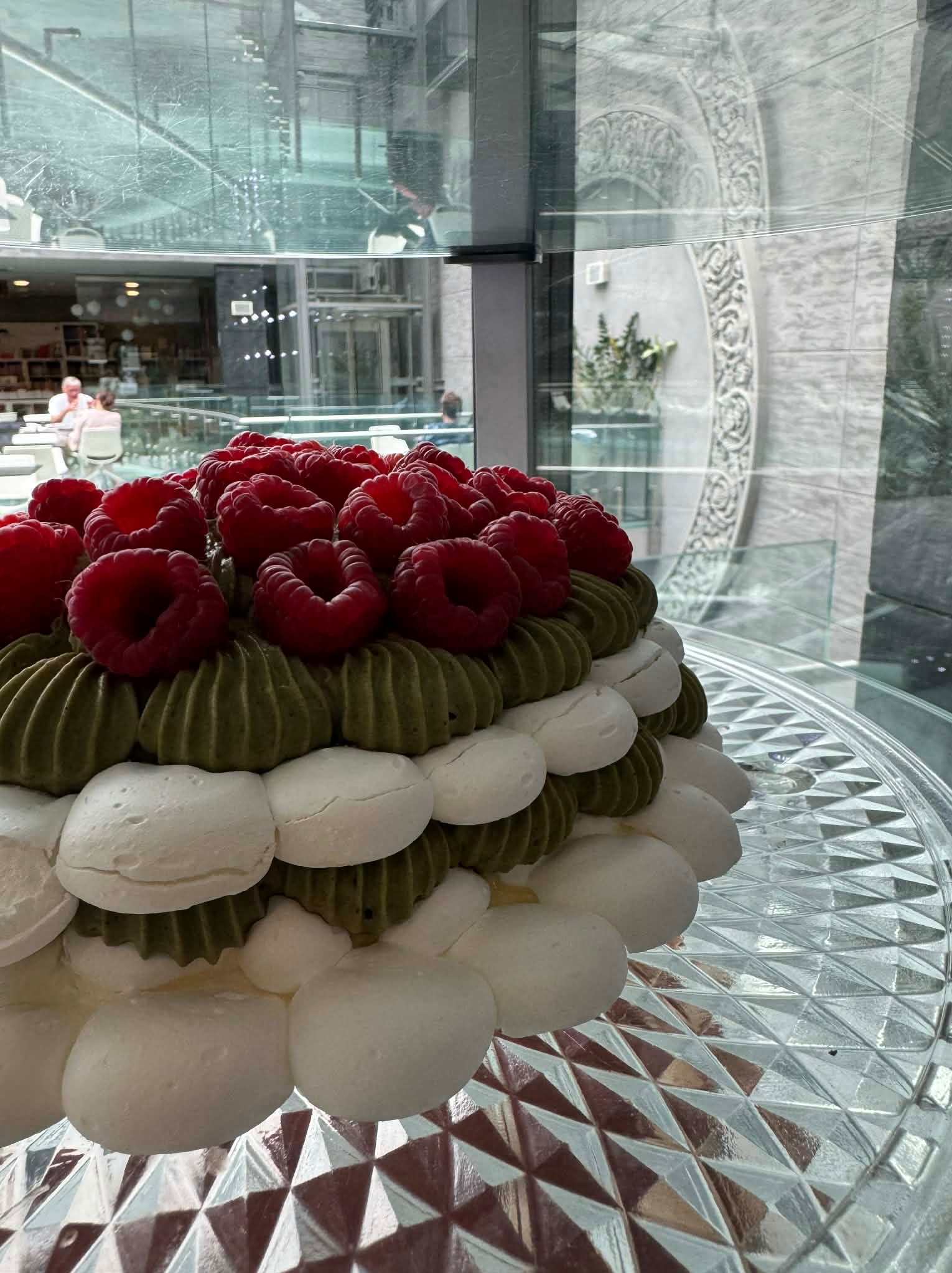 Elegancki tort z malinami, zielonym kremem i białymi bezami na szklanym paterze, na tle nowoczesnego wnętrza z dużymi oknami.