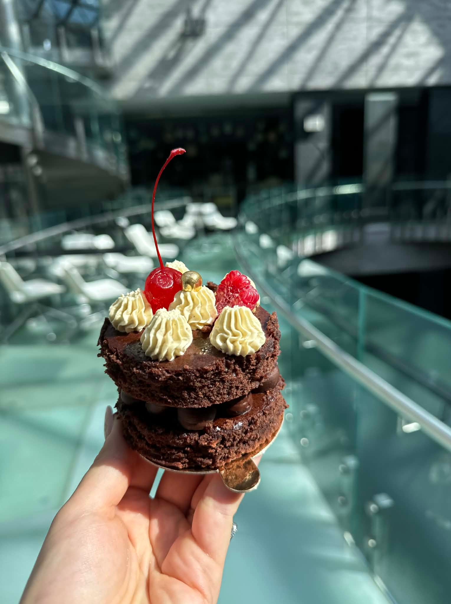 Dwupoziomowe, czekoladowe ciastko z kremem, maliną i wisienką na tle nowoczesnego wnętrza. Delikatne światło podkreśla detale deseru.