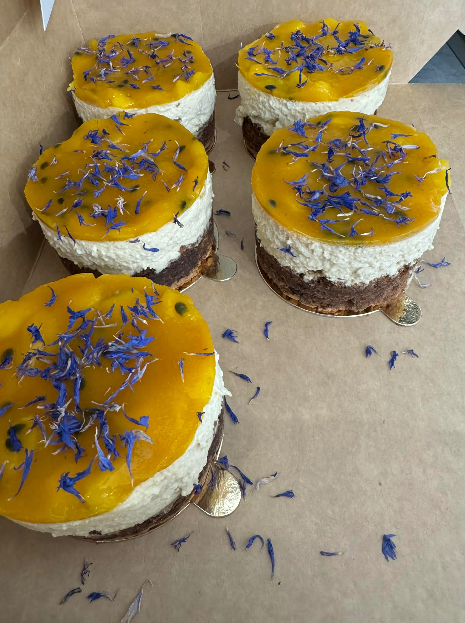 Cztery mini torty z musem, galaretką z marakui i dekoracją z niebieskich kwiatów w kartonowym pudełku. Widok z góry, delikatne światło.