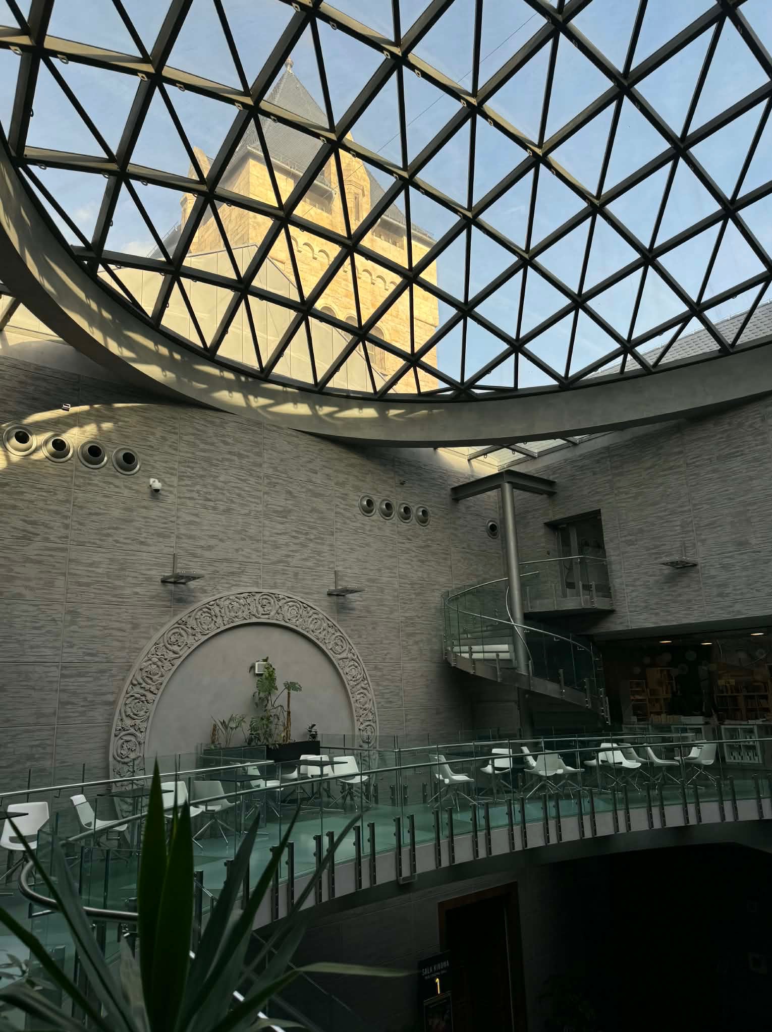 Nowoczesne wnętrze z przeszklonym dachem w kształcie kopuły, widoczne krzesła i stoliki, spiralne schody oraz dekoracyjny element na ścianie.