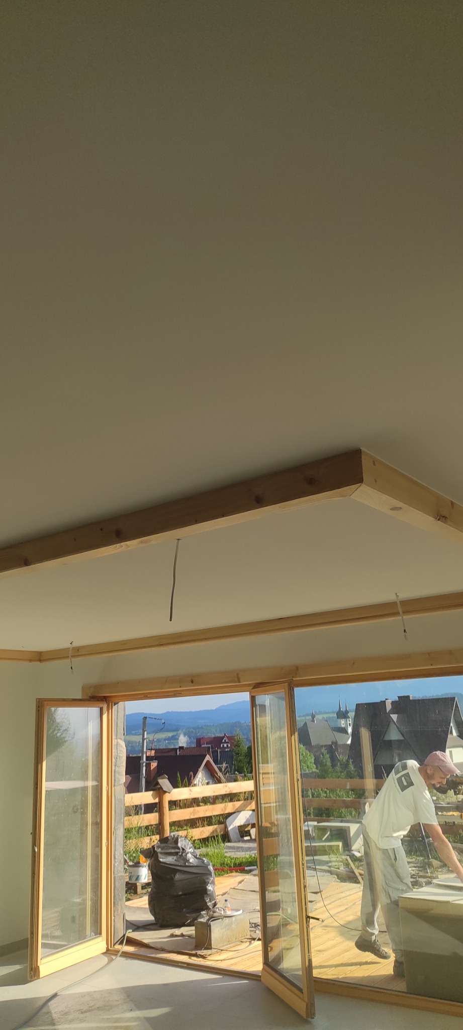 Prace wykończeniowe w jasnym pokoju z dużymi oknami i widokiem na góry. Widoczny pracownik podczas szpachlowania sufitu. Elementy drewniane w konstrukcji sufitu.