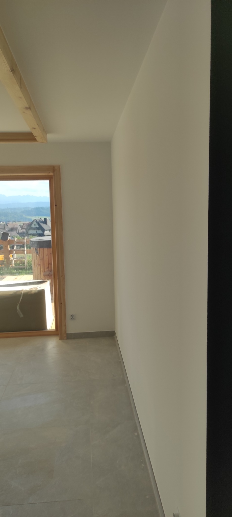 Jasne wnętrze po remoncie: gładkie, białe ściany, szara podłoga z płytek, drewniane wykończenie okna z widokiem na góry. Minimalistyczny design, świeżo po malowaniu.