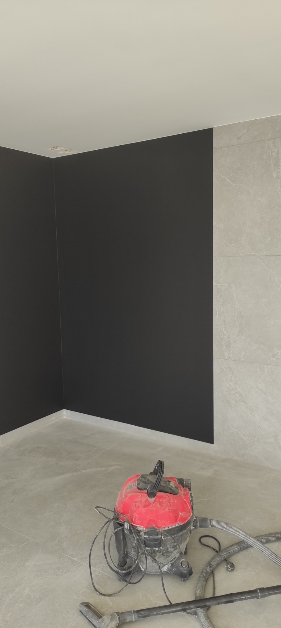 Wnętrze po malowaniu ścian na czarno, z fragmentem ściany wyłożonej płytkami. Na podłodze widoczny odkurzacz przemysłowy, sugerujący prace porządkowe po malowaniu.