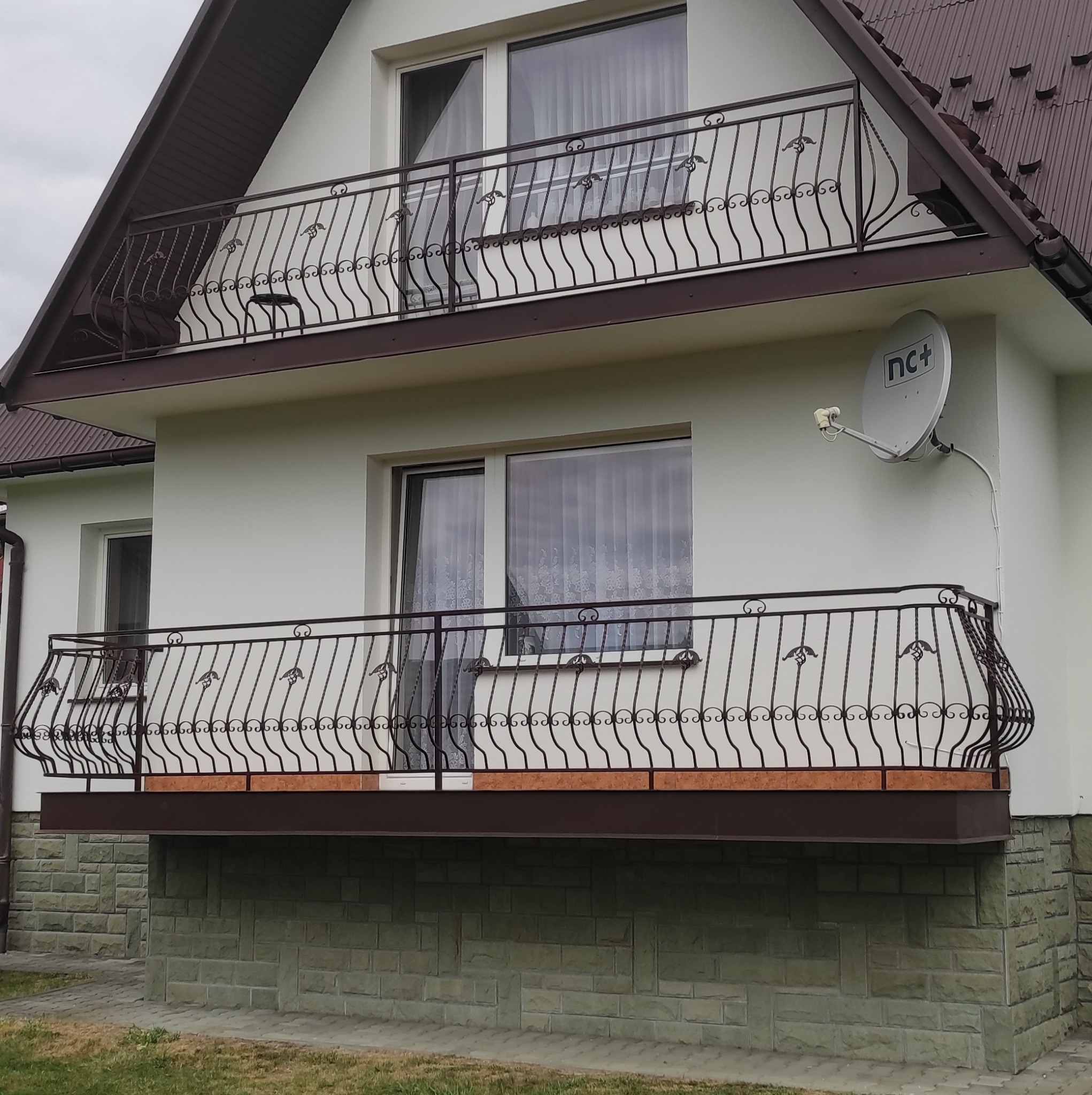 Elewacja domu z balkonami o zdobionych, metalowych balustradach. Widoczna antena satelitarna na ścianie. Dolna część elewacji wykończona kamieniem.
