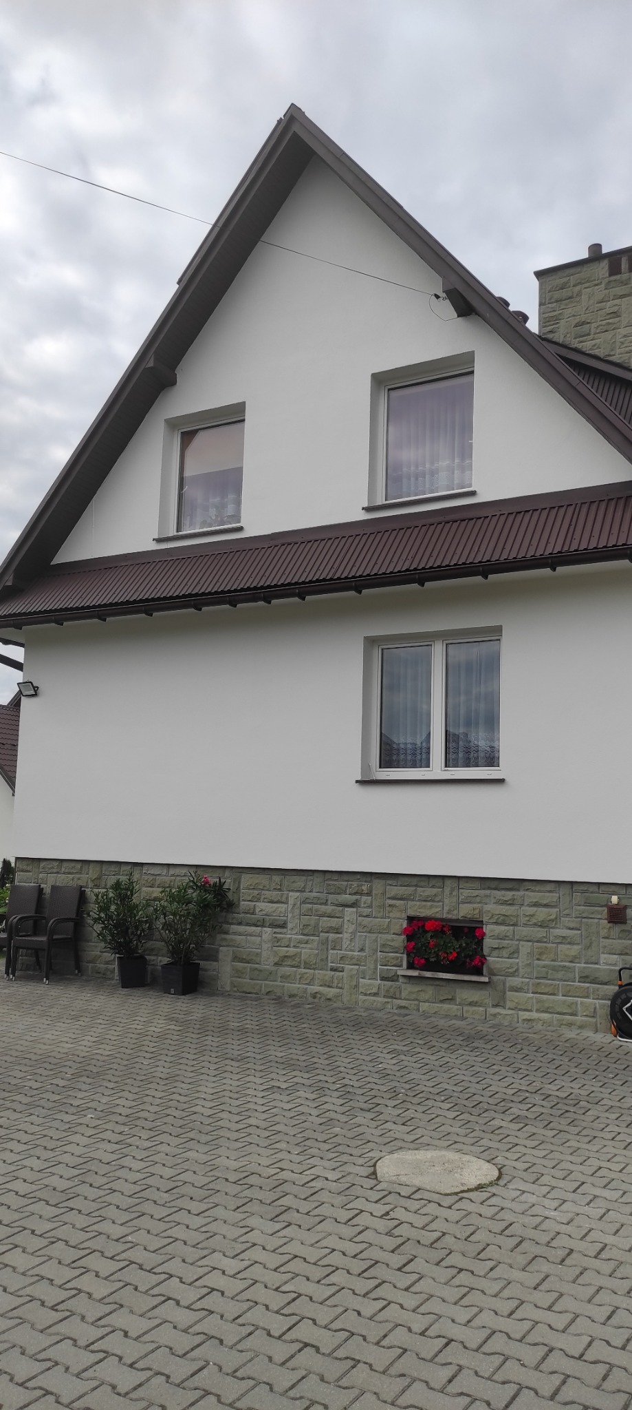 Biała elewacja domu z brązowym dachem w Krościenku nad Dunajcem. Kamienna podmurówka z kwiatami, okna z firankami. Widok na fragment posesji.