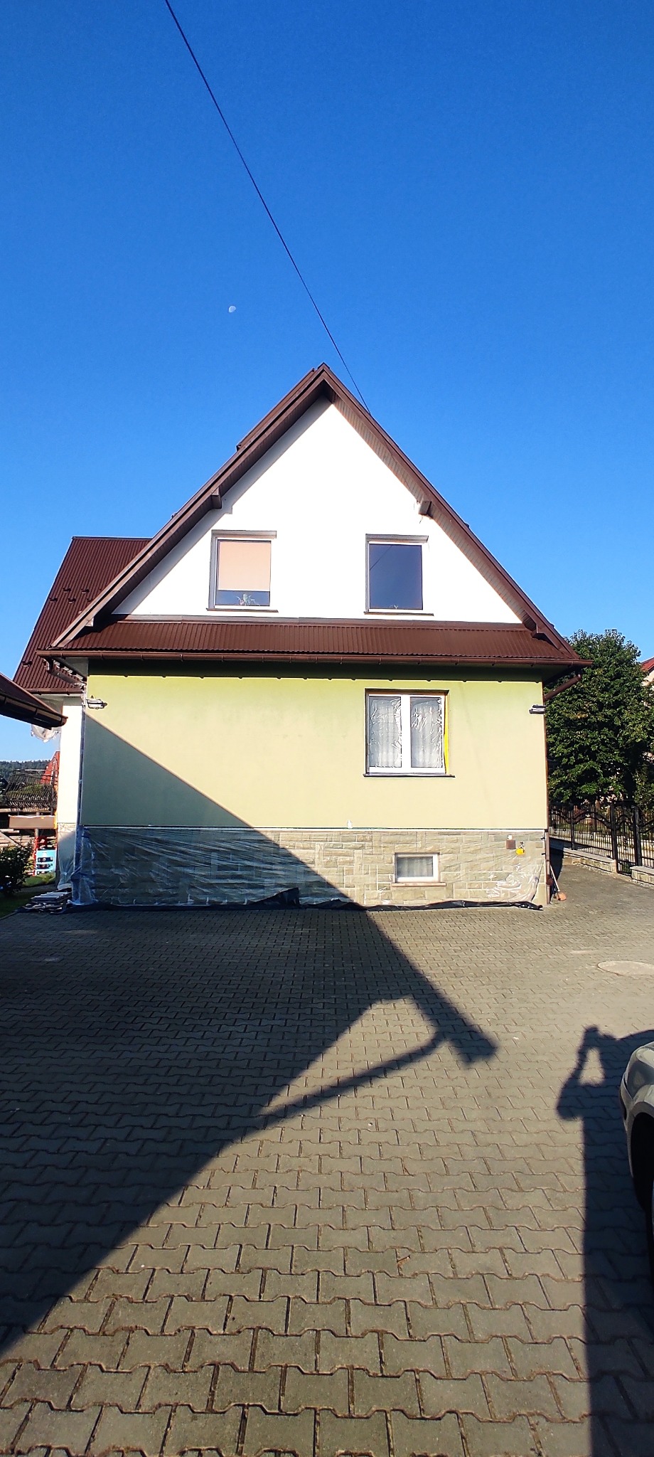 Dwukondygnacyjny dom z zieloną elewacją, zabezpieczony folią malarską. Widoczne okna, brązowy dach i kostka brukowa. Błękitne niebo w tle.