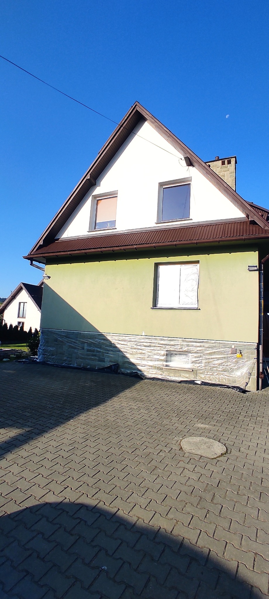 Dom jednorodzinny w trakcie prac elewacyjnych: okna zabezpieczone folią, świeżo pomalowana ściana w odcieniach zieleni, widoczne rusztowania i kostka brukowa.