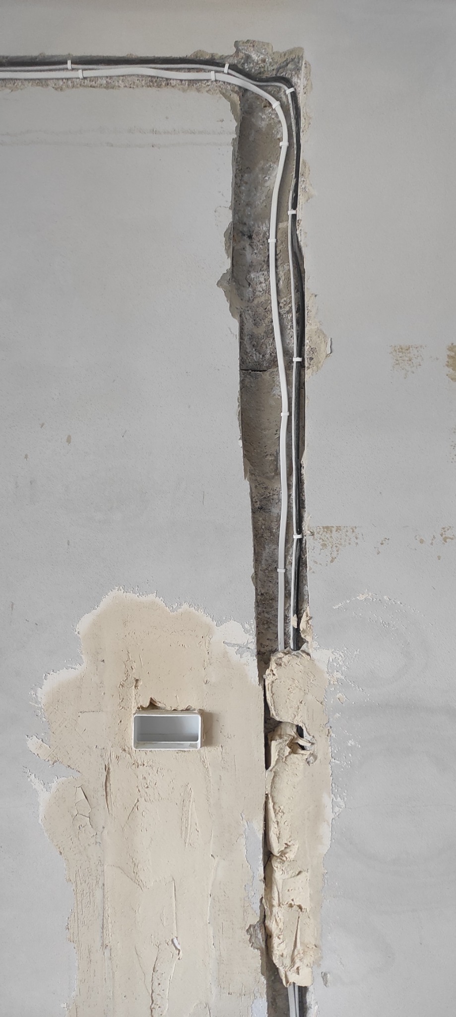 Bruzdy z przewodami elektrycznymi w ścianie przed szpachlowaniem. Widoczne kable przymocowane klamrami. Fragment ściany z nałożoną warstwą szpachli i puszką elektryczną.