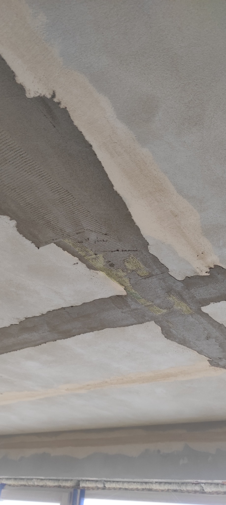Sufit po szpachlowaniu z widoczną siatką wzmacniającą na łączeniach płyt. Szpachla w odcieniach szarości i bieli. Widok z dołu, fragment okna w dolnej części kadru.