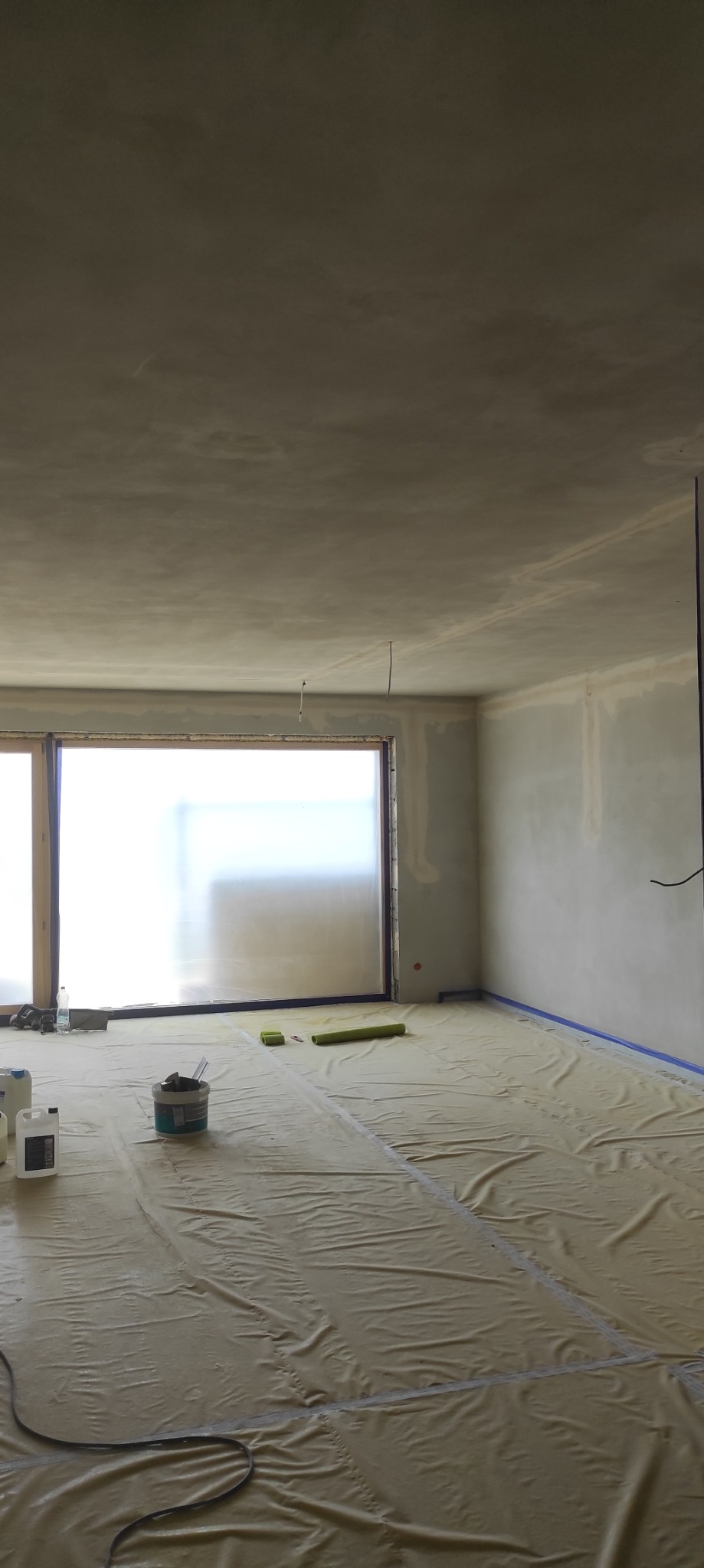 Pokój w trakcie prac wykończeniowych: zabezpieczona podłoga folią, ściany i sufit po szpachlowaniu, widoczne narzędzia i materiały malarskie. Duże okno z osłoną.