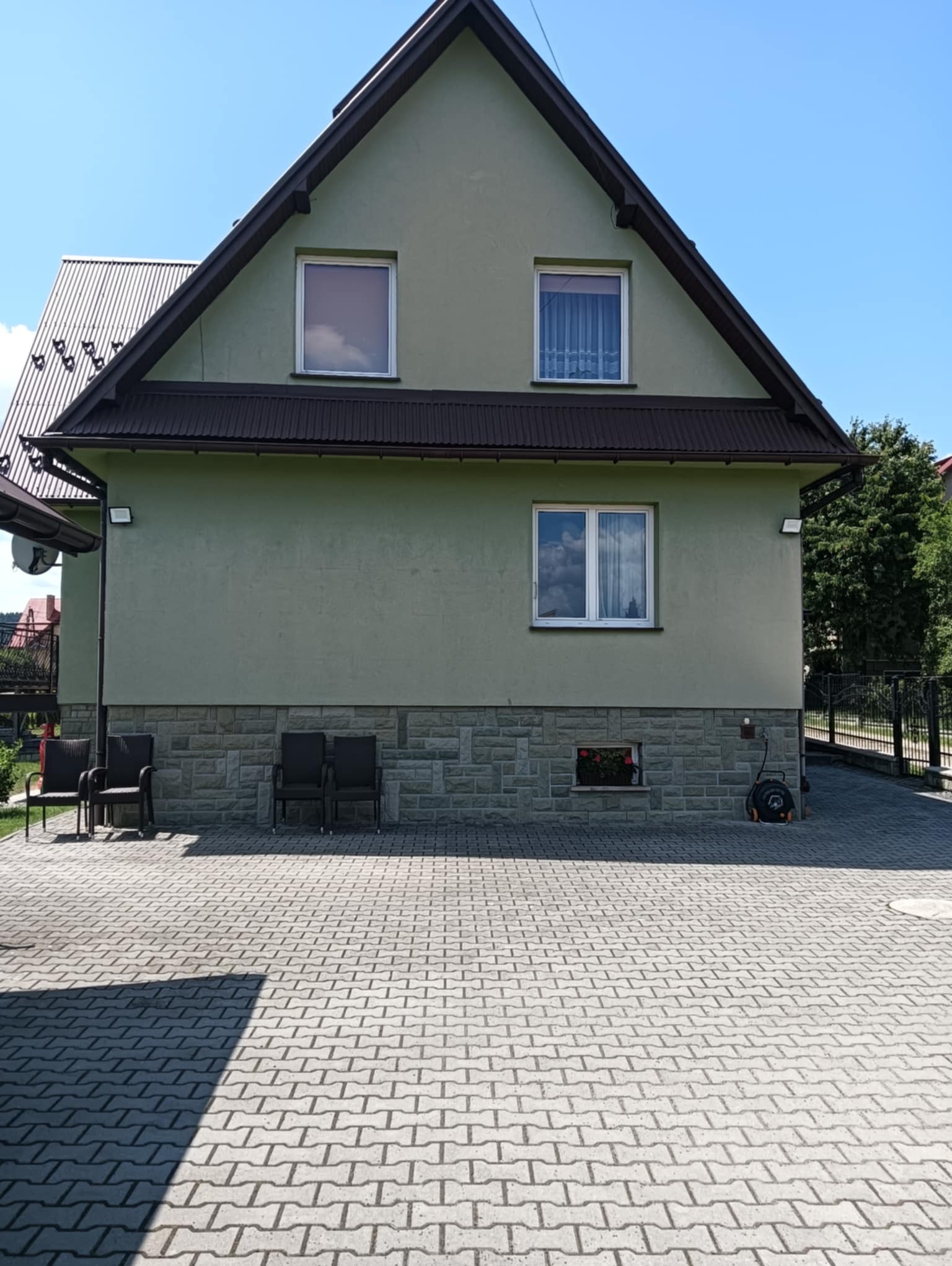 Dwukondygnacyjny dom z zieloną elewacją i brązowym dachem, z widocznym fragmentem kostki brukowej i trzema krzesłami na zewnątrz. Widok z przodu, w słoneczny dzień.