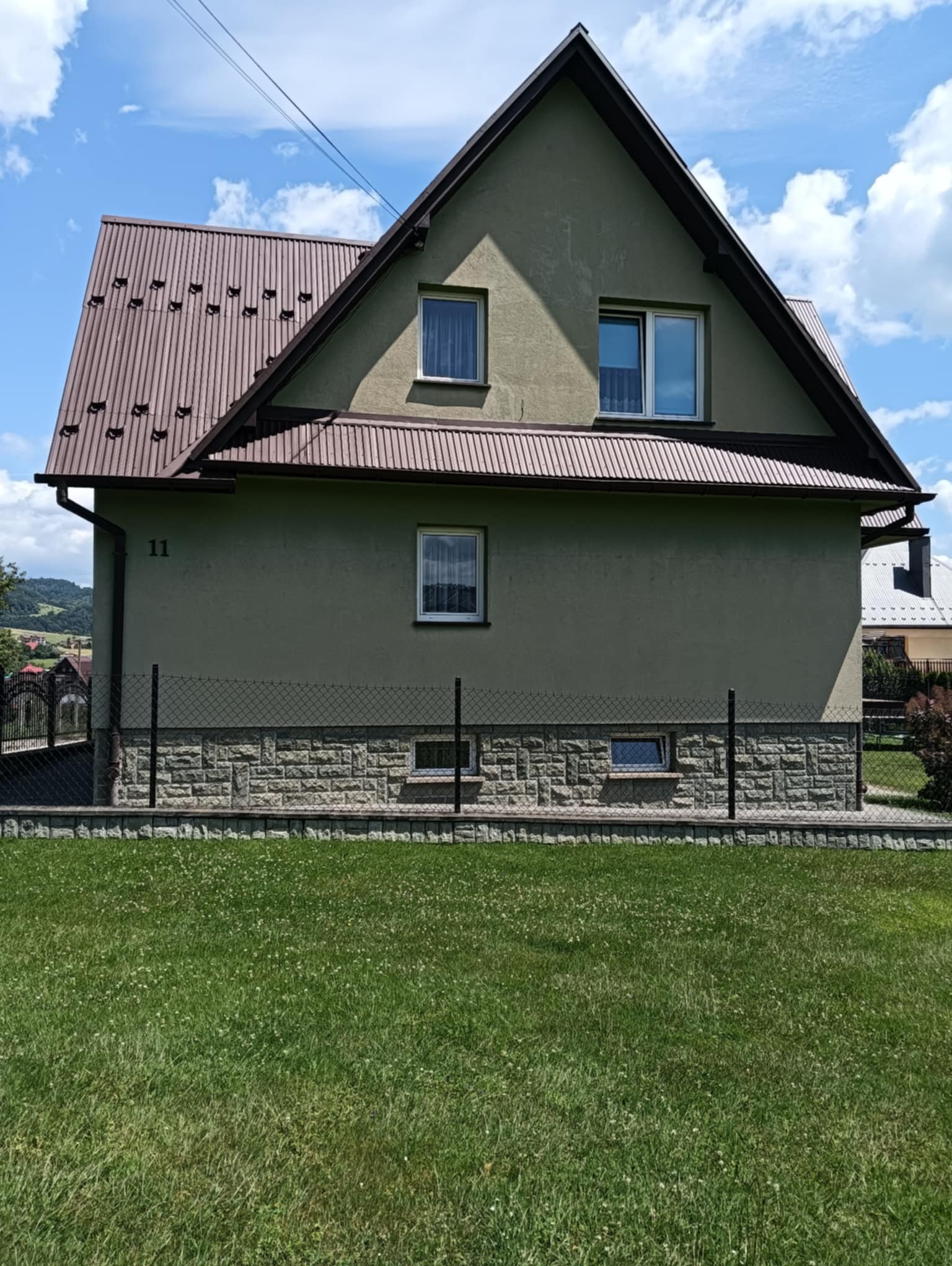 Dom jednorodzinny z brązowym dachem i elewacją w odcieniach zieleni, numer 11, ogrodzony siatką, z widocznym trawnikiem i fragmentem krajobrazu w tle.