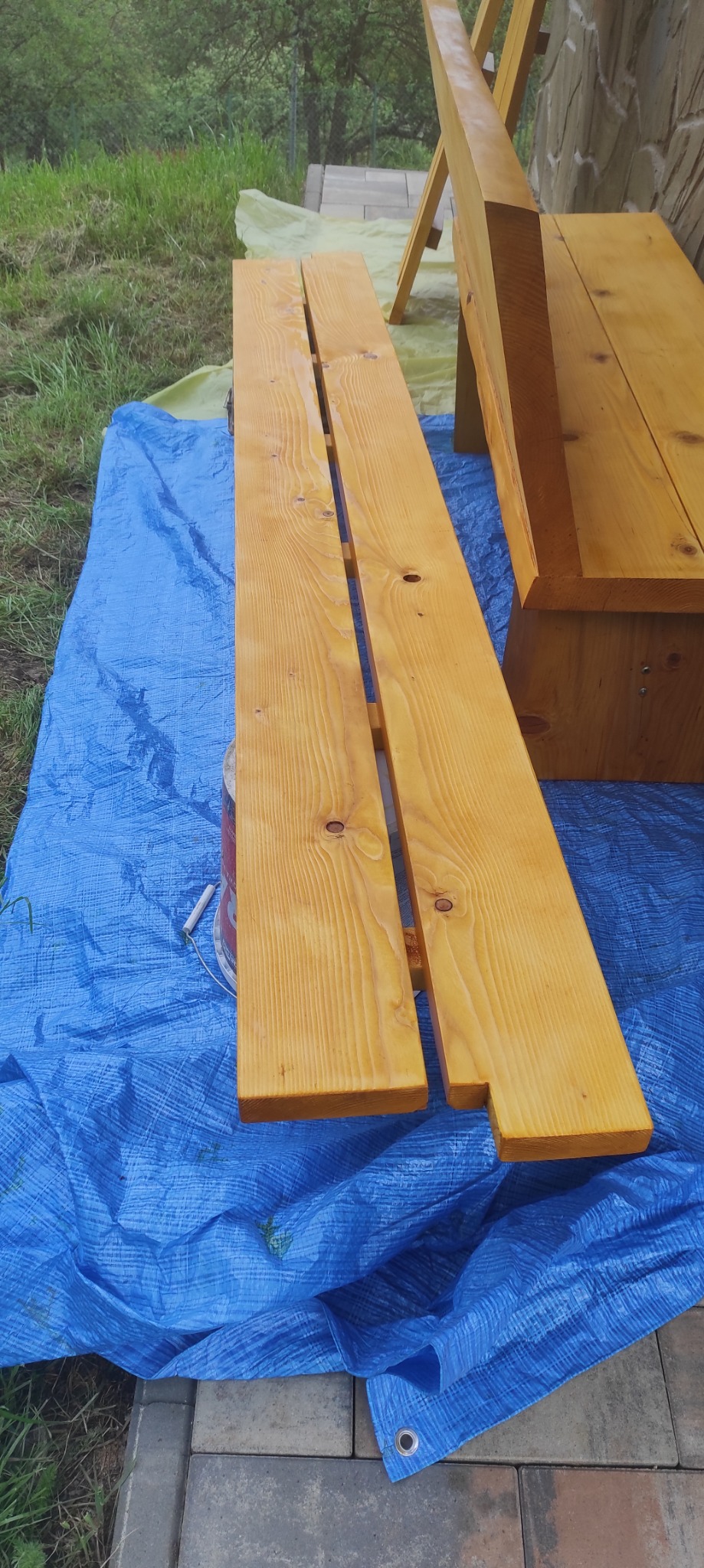 Impregnowane drewniane elementy ławki ogrodowej na niebieskiej folii ochronnej. Widoczna puszka z impregnatem i pędzel. Trawa i kamienna ściana w tle.