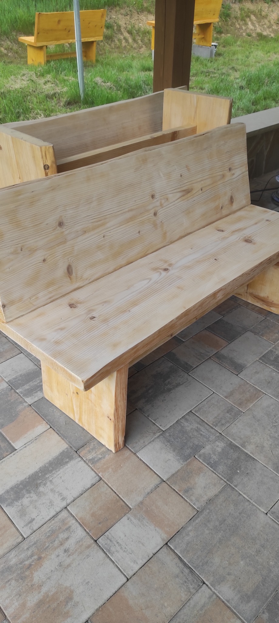Drewniana ławka ogrodowa z oparciem, wykonana z jasnego drewna, ustawiona na kostce brukowej. W tle widoczne inne elementy drewniane i zieleń.