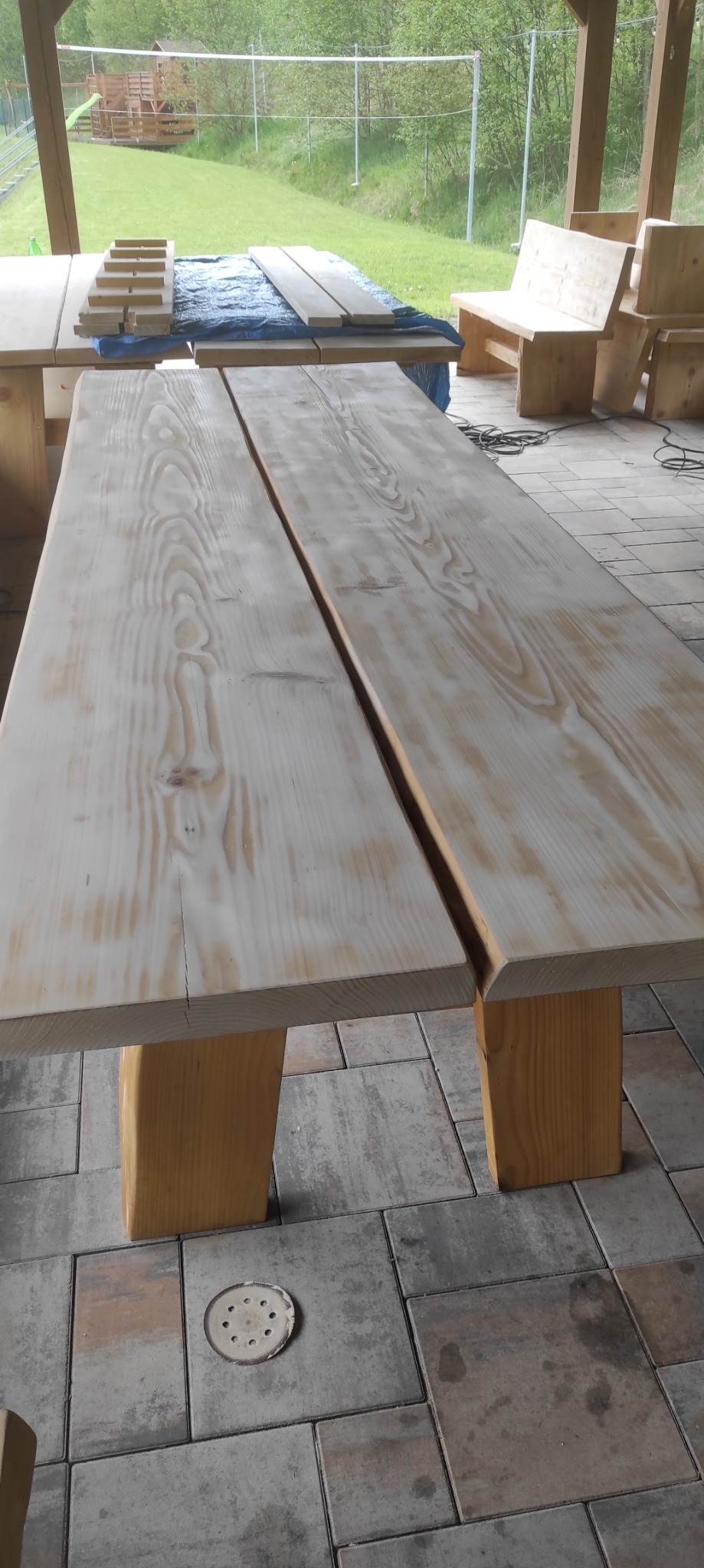 Długi, drewniany stół z dwoma blatami i grubymi nogami, stojący na kostce brukowej. W tle widać inne meble ogrodowe i zieleń. Na podłodze leży krążek ścierny.