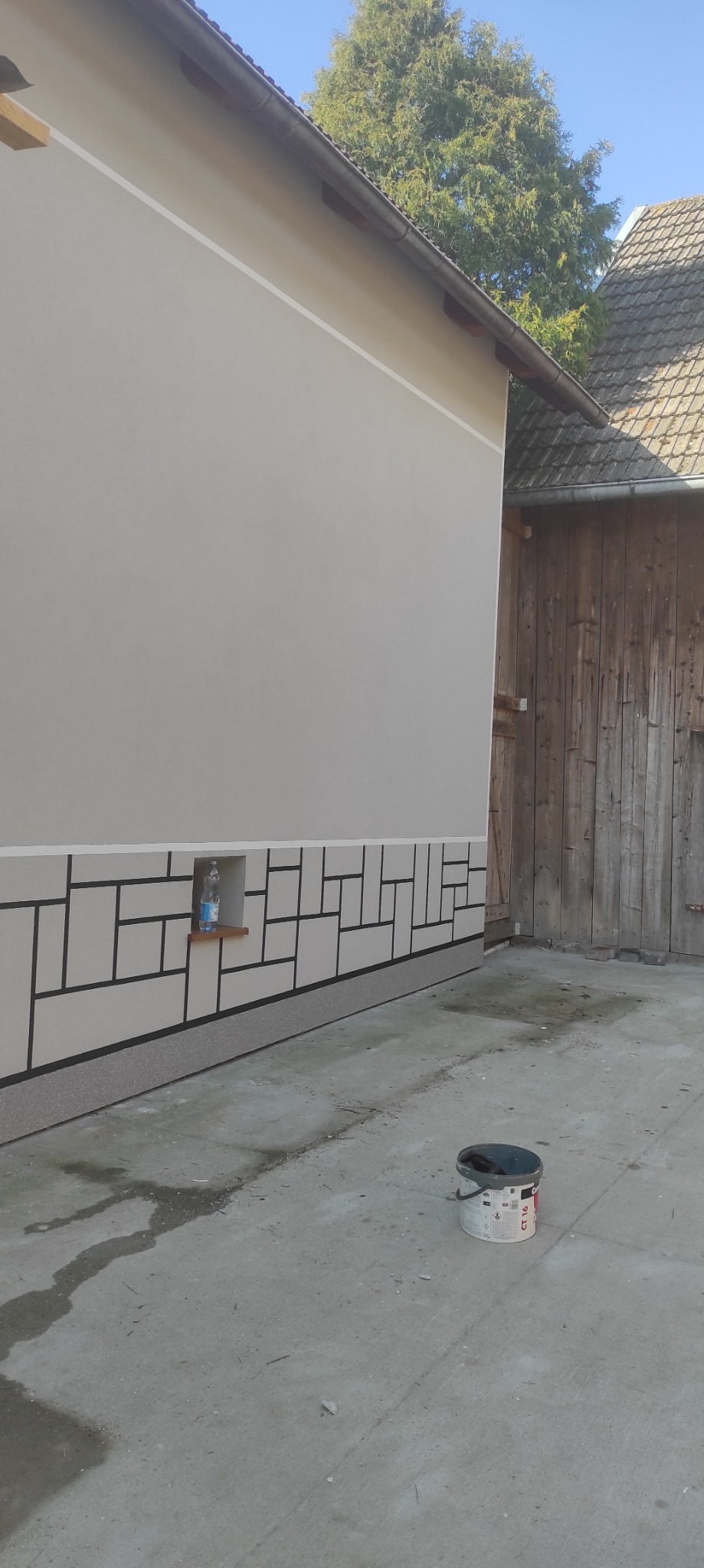 Elewacja budynku po malowaniu z dekoracyjnym wzorem geometrycznym w dolnej części. Wiadro z farbą na posadzce. Widoczny fragment drewnianej stodoły.