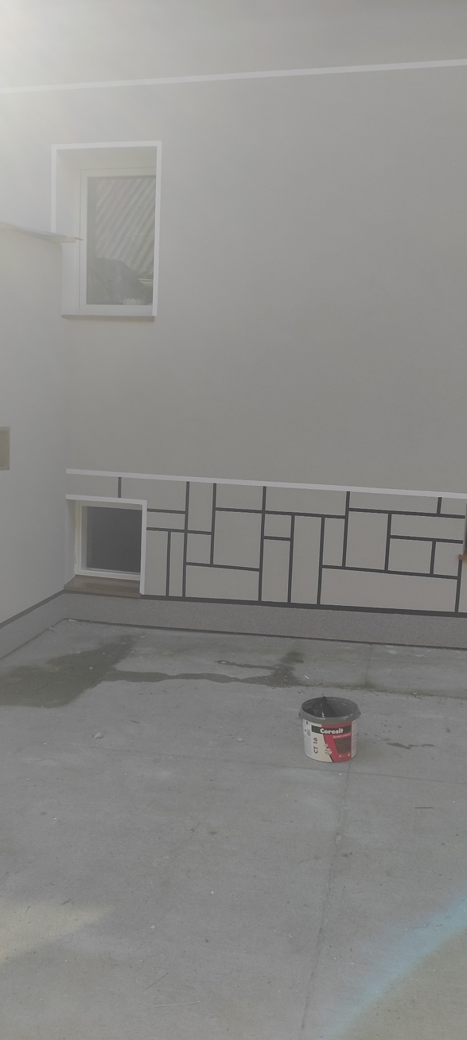 Ściana z geometrycznym wzorem w stylu Mondriana, pomalowana na szaro z czarnymi liniami. Widoczny fragment okna i wiadro z farbą na podłodze.