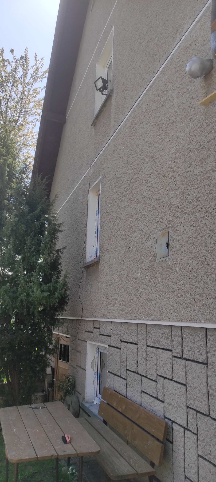 Elewacja domu z widocznymi oknami przed wymianą, fragmentem ocieplenia i lampą zewnętrzną. Widoczny stół i ławka.