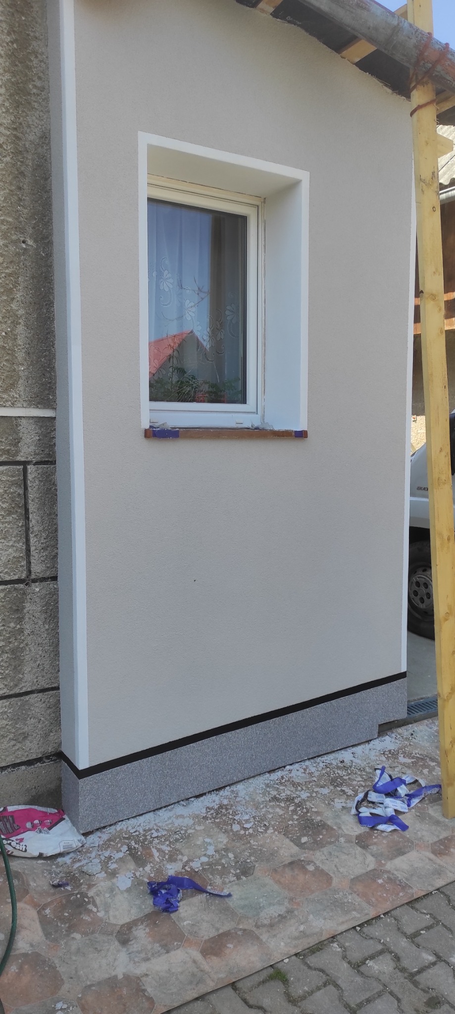 Fragment elewacji budynku po remoncie, z oknem i świeżo pomalowaną ścianą w odcieniach beżu. Widoczne ślady prac na podłożu i fragment rusztowania.