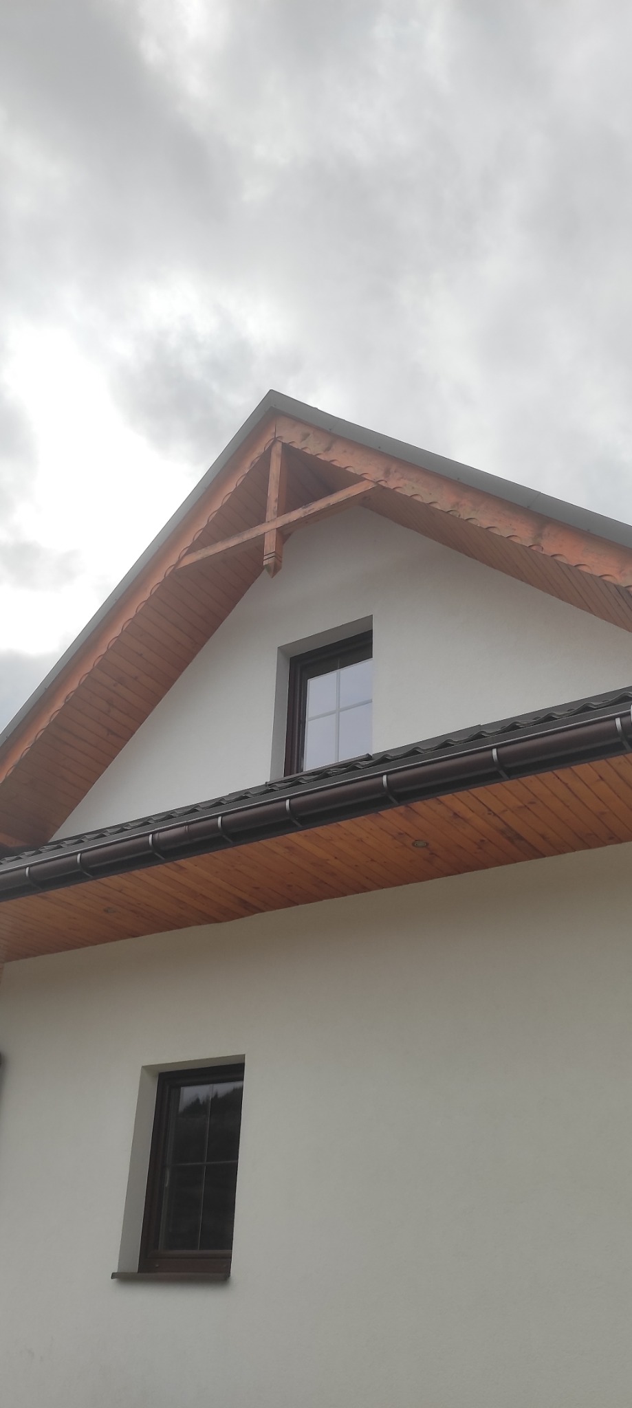 Biała elewacja domu z drewnianym wykończeniem dachu i okien. Widoczne dwa okna, jedno na poddaszu. Pochmurne niebo w tle. Detale drewniane z dekoracyjnymi wycięciami.