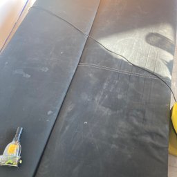 Król Kanapy - Ciemna, dwuczęściowa kanapa z widocznymi plamami, częściowo czyszczona odkurzaczem piorącym. Widoczne ślady po czyszczeniu. Oświetlenie dzienne.