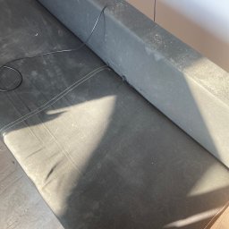 Król Kanapy - Ciemna, zabrudzona sofa z widocznymi plamami i kurzem na tapicerce. Widoczne ślady użytkowania i zarysowania. Kabel zwisający z boku sofy.