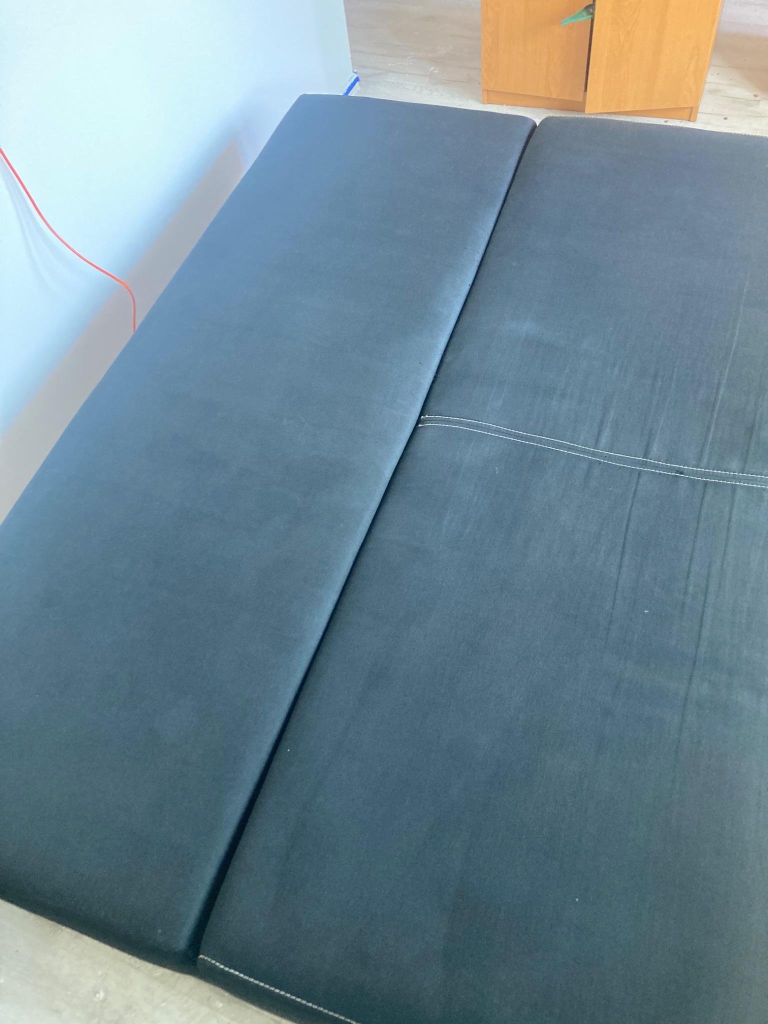 Ciemnoszara, rozłożona kanapa z widocznym szwem, ustawiona przy białej ścianie i drewnianej szafce. Ujęcie z góry, prezentujące czystą tapicerkę.