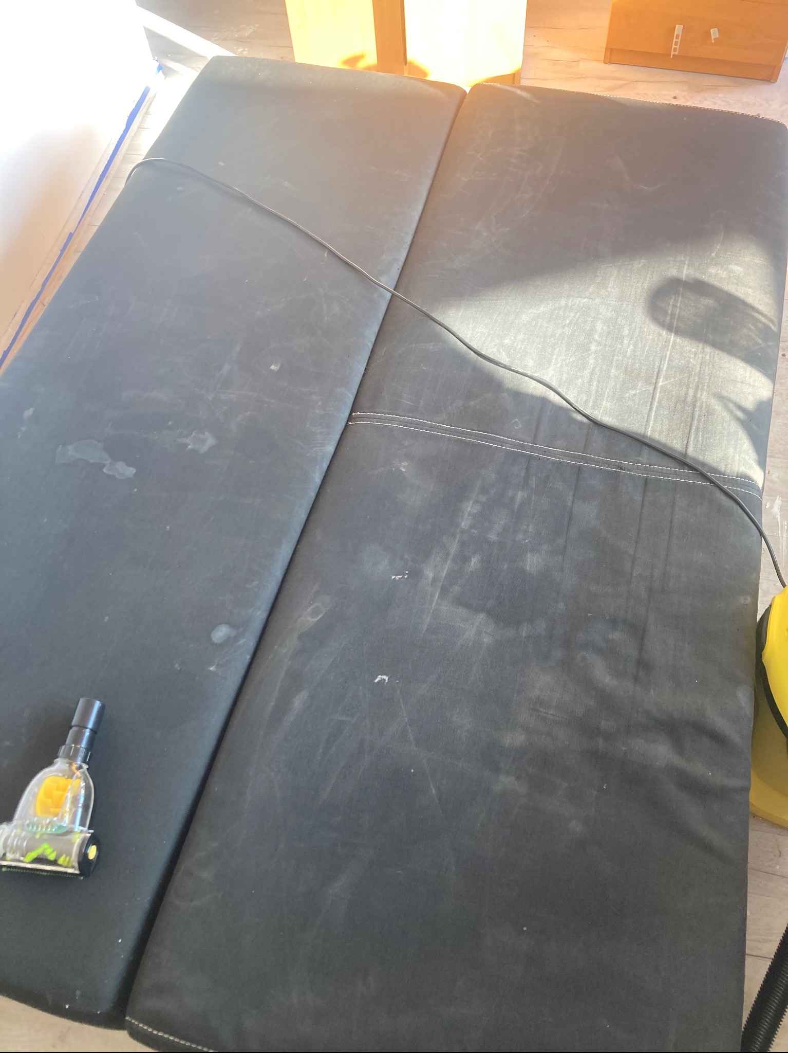 Ciemna, dwuczęściowa kanapa z widocznymi plamami, częściowo czyszczona odkurzaczem piorącym. Widoczne ślady po czyszczeniu. Oświetlenie dzienne.