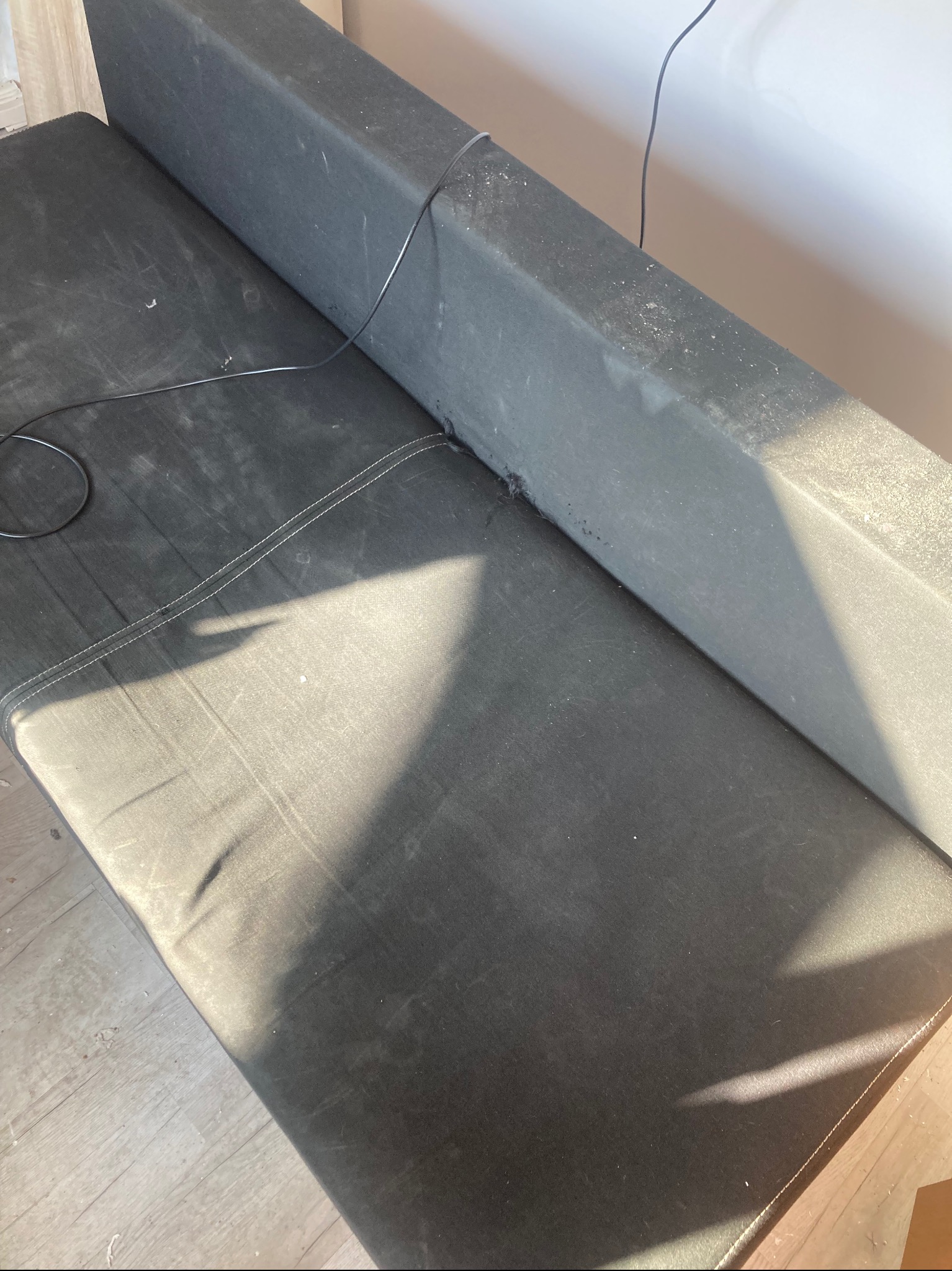 Ciemna, zabrudzona sofa z widocznymi plamami i kurzem na tapicerce. Widoczne ślady użytkowania i zarysowania. Kabel zwisający z boku sofy.