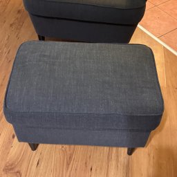 EDISON PRO CLEAN - Granatowy podnóżek tapicerowany tkaniną, ustawiony na drewnianej podłodze, z fragmentem fotela w tle. Widoczne subtelne detale faktury materiału.