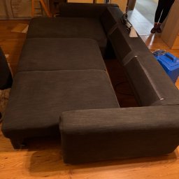 EDISON PRO CLEAN - Ciemnoszara, narożna kanapa z widocznymi śladami użytkowania, częściowo czyszczona. W tle widoczny sprzęt do czyszczenia tapicerki i elementy wyposażenia wnętrza.