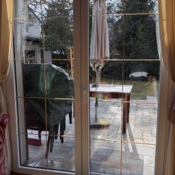 EDISON PRO CLEAN - Białe okno balkonowe z widokiem na taras i ogród. Na podłodze wiadro z akcesoriami do mycia okien i czarny ręcznik. Słoneczny dzień.