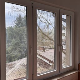 EDISON PRO CLEAN - Trzyczęściowe okno PCV z widokiem na zewnątrz: drzewa iglaste, budynki i dach pokryty liśćmi. Wnętrze pokoju z komodą i lampą.