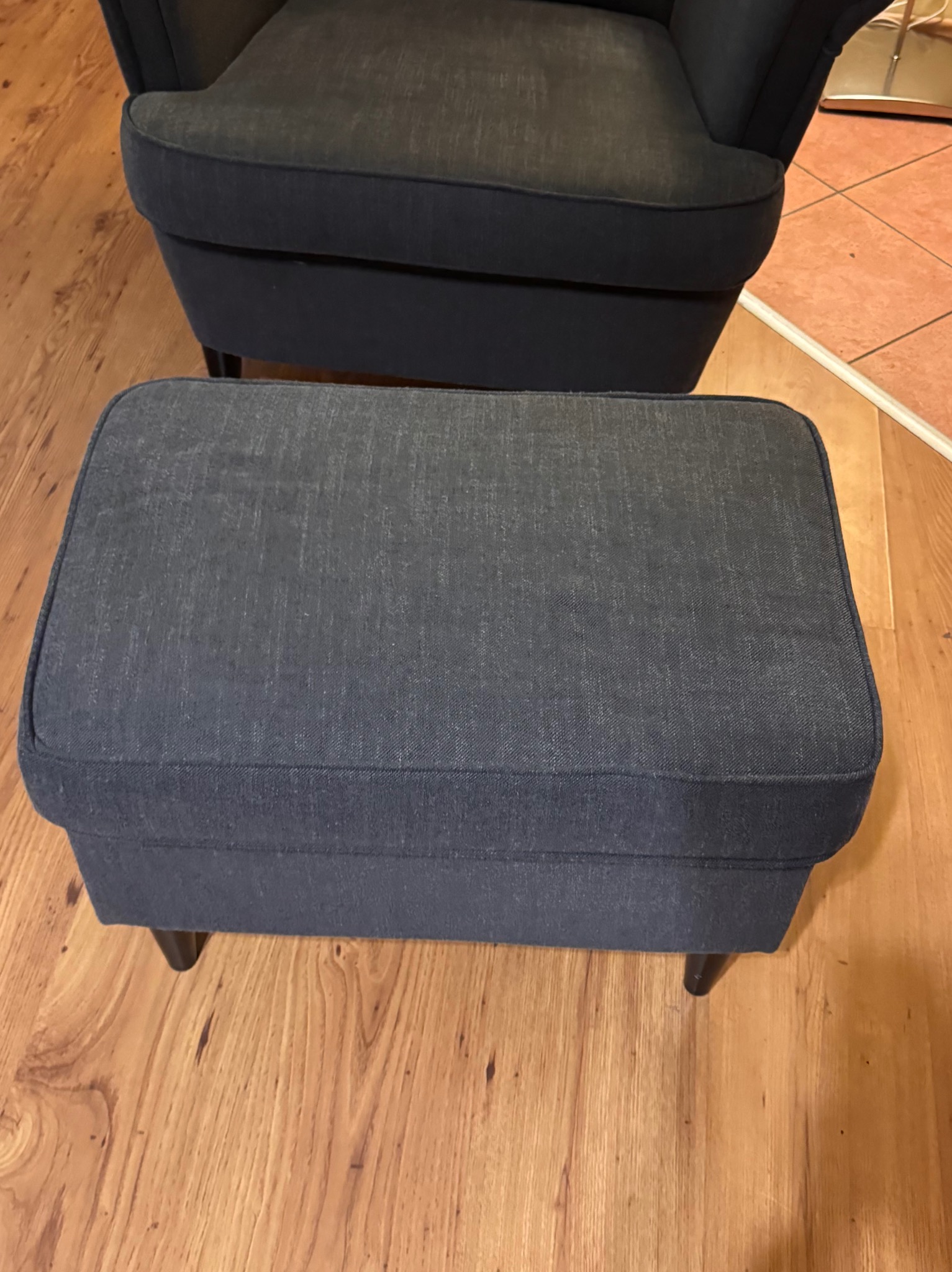 Granatowy podnóżek tapicerowany tkaniną, ustawiony na drewnianej podłodze, z fragmentem fotela w tle. Widoczne subtelne detale faktury materiału.