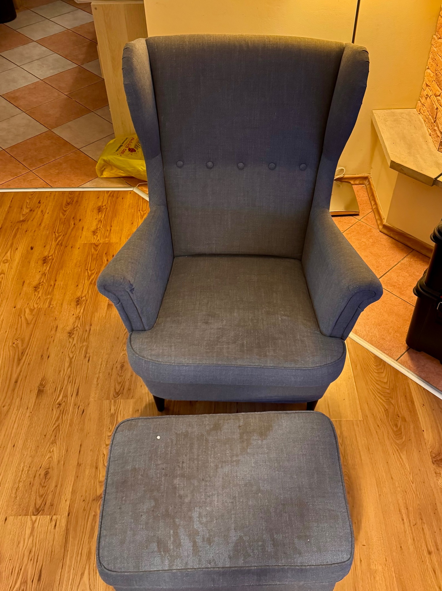 Szary fotel typu uszak z podnóżkiem na drewnianej podłodze. Widoczne plamy na podnóżku. W tle fragmenty pomieszczenia z różnymi rodzajami podłóg.