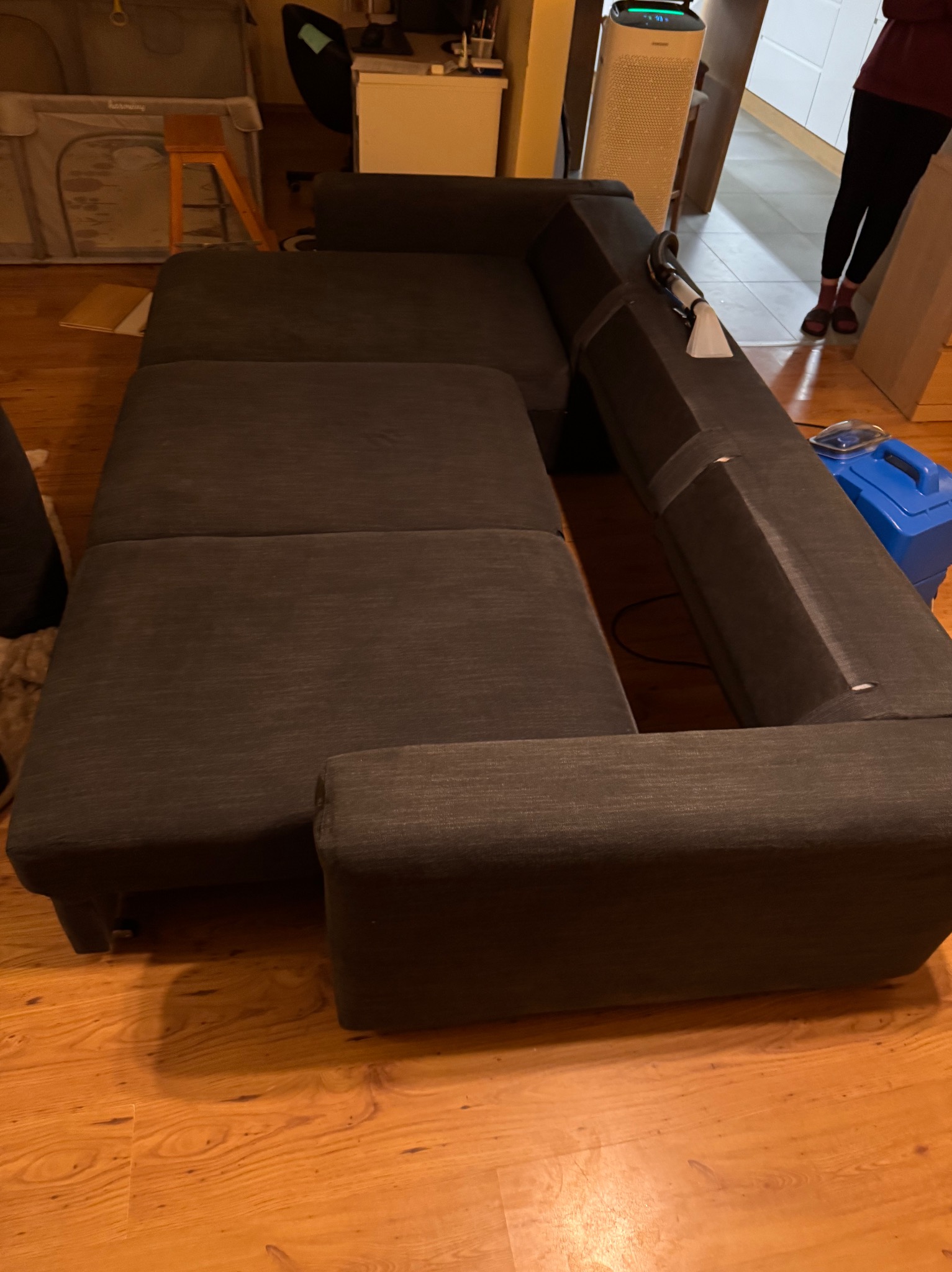 Ciemnoszara, narożna kanapa z widocznymi śladami użytkowania, częściowo czyszczona. W tle widoczny sprzęt do czyszczenia tapicerki i elementy wyposażenia wnętrza.