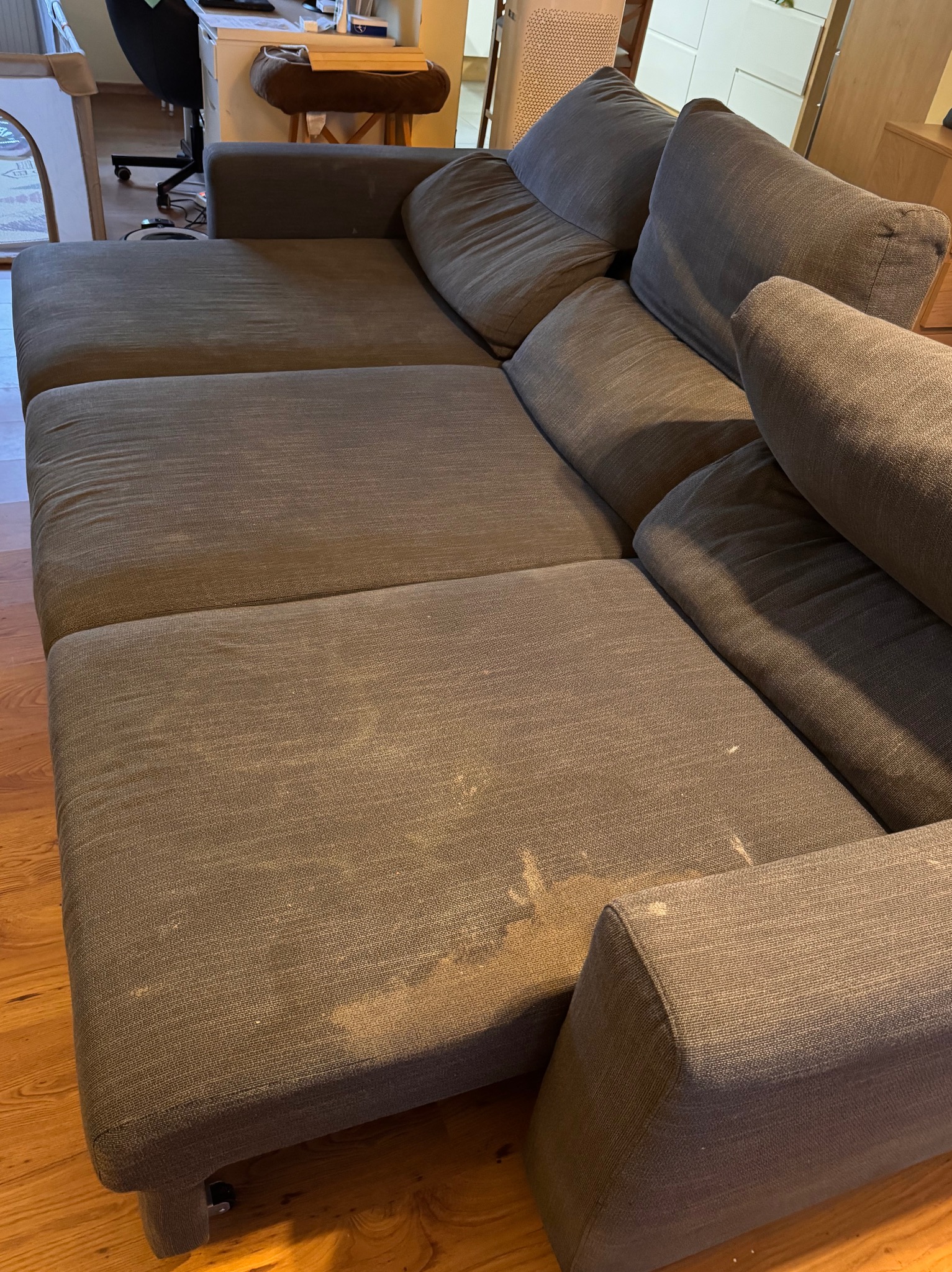 Szary narożnik z widocznymi plamami na siedzisku, ustawiony na drewnianej podłodze. Widoczne poduszki i fragmenty mebli w tle. Potrzeba odświeżenia tapicerki.