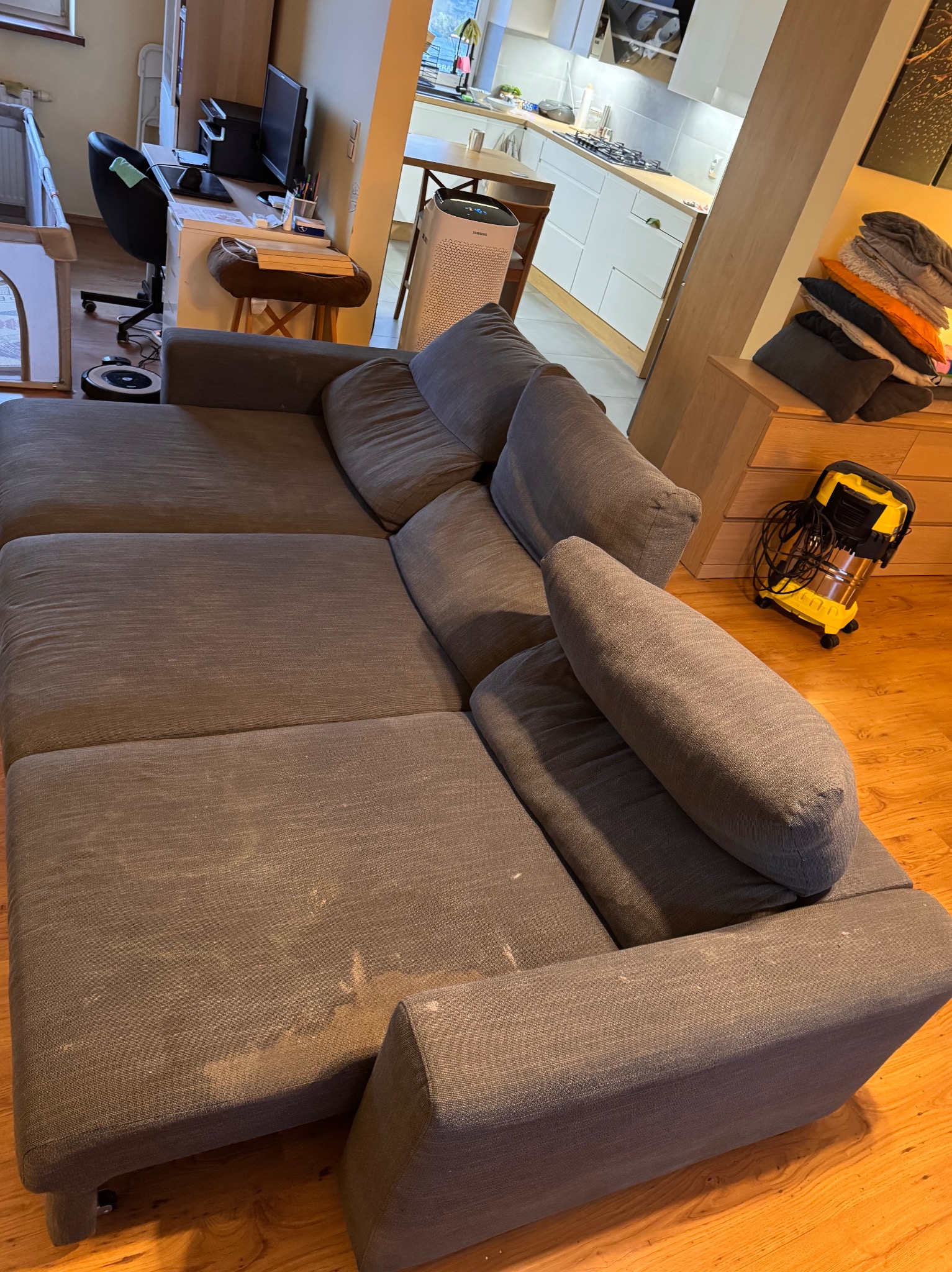 Szara, modułowa kanapa z widocznymi plamami na siedzisku i podłokietniku, obok żółty odkurzacz piorący. W tle widoczny fragment mieszkania.