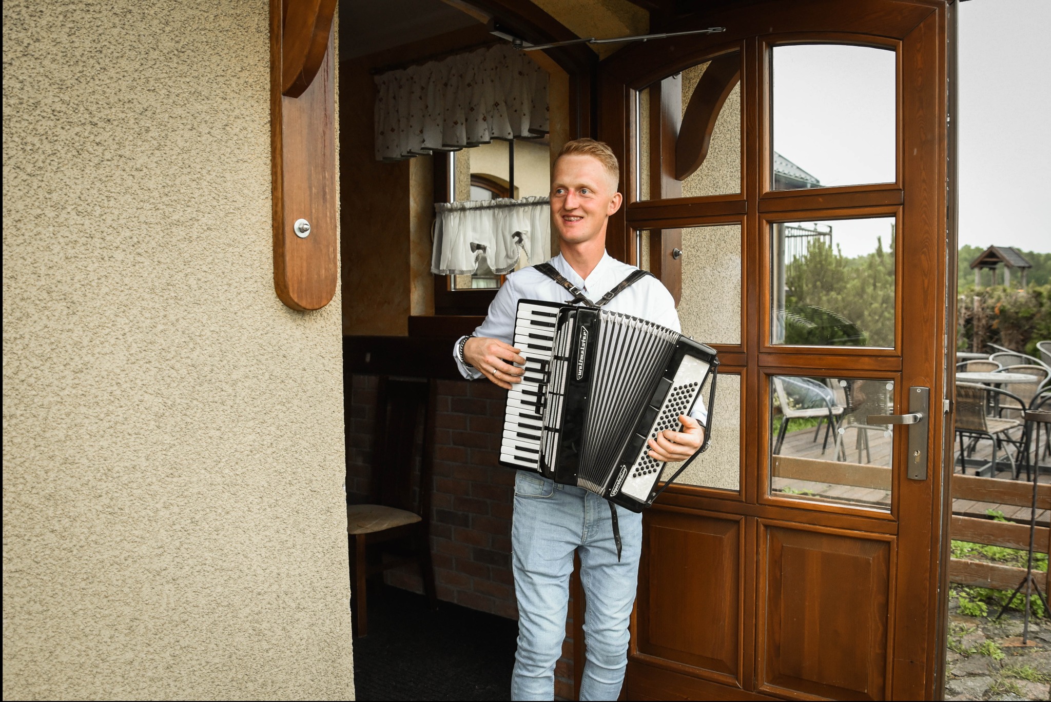 Młody akordeonista w jasnej koszuli i jeansach, grający na akordeonie przed wejściem do budynku z drewnianymi drzwiami i widokiem na ogród.