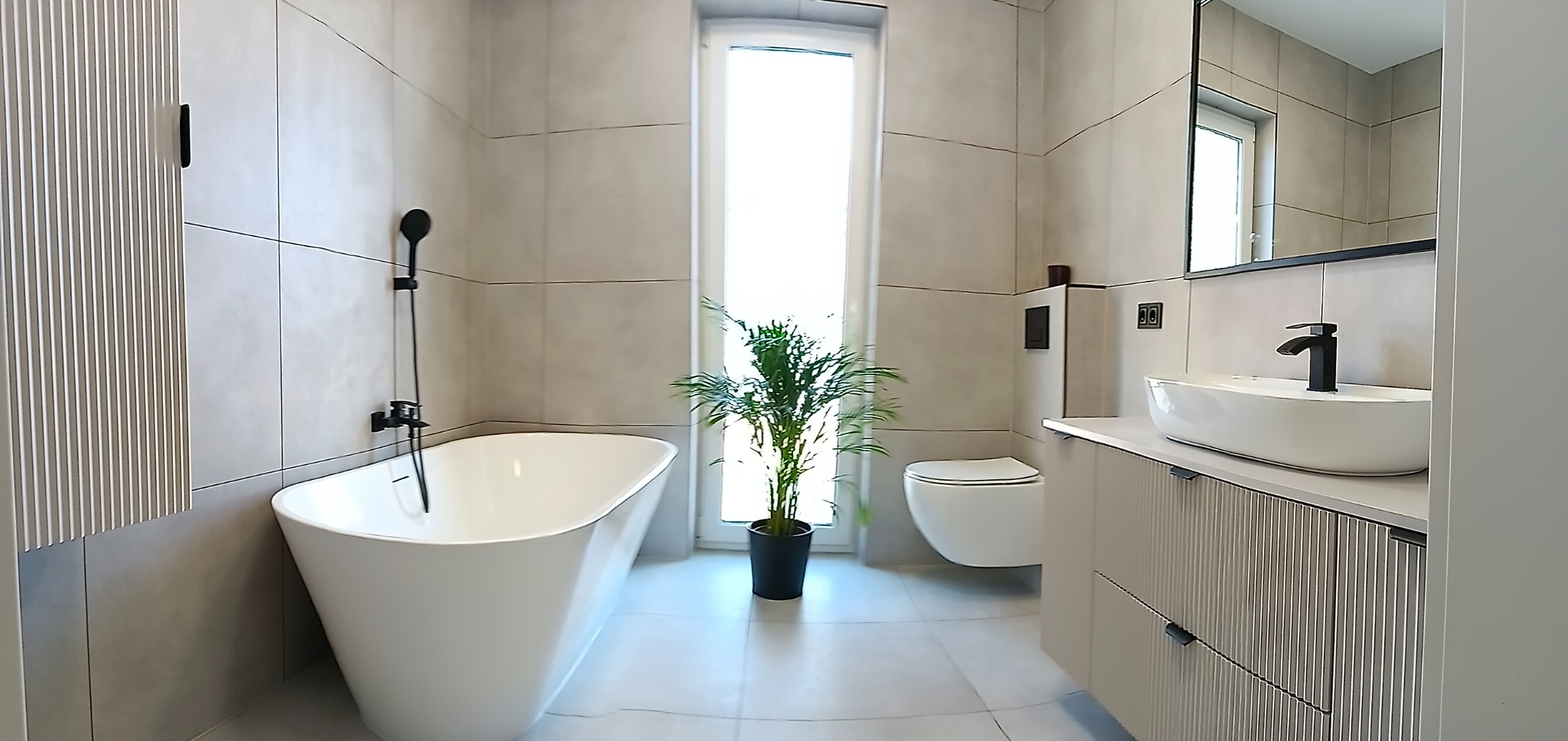 Nowoczesna łazienka w beżowych płytkach z wolnostojącą wanną, umywalką nablatową i rośliną doniczkową przy oknie. Minimalistyczny design z czarnymi akcentami.