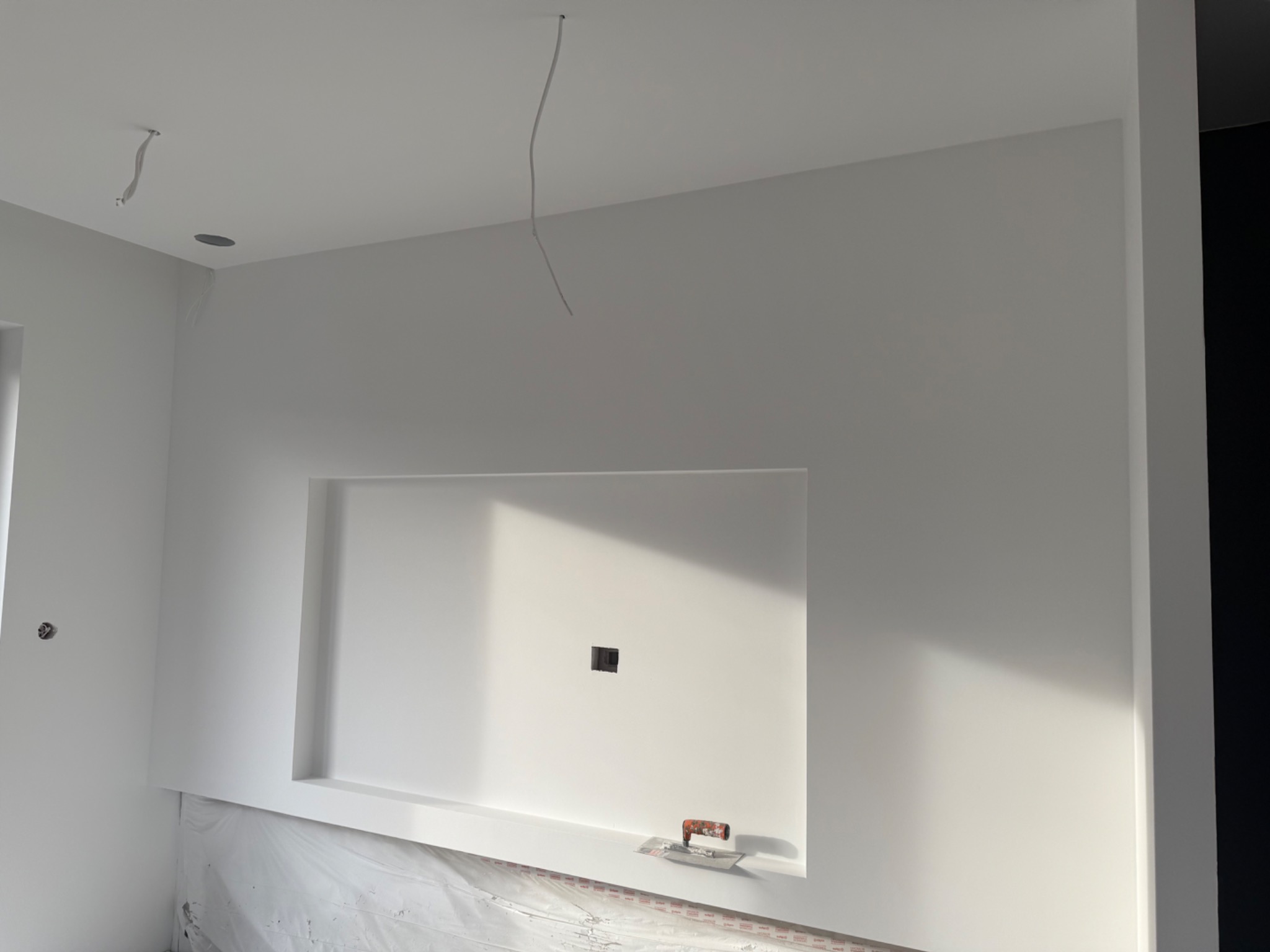 Biała ściana z wnęką pod telewizor po szpachlowaniu, widoczne narzędzie (szpachelka) na dolnej krawędzi wnęki. Kabel zwisa z sufitu. Folia ochronna na podłodze.