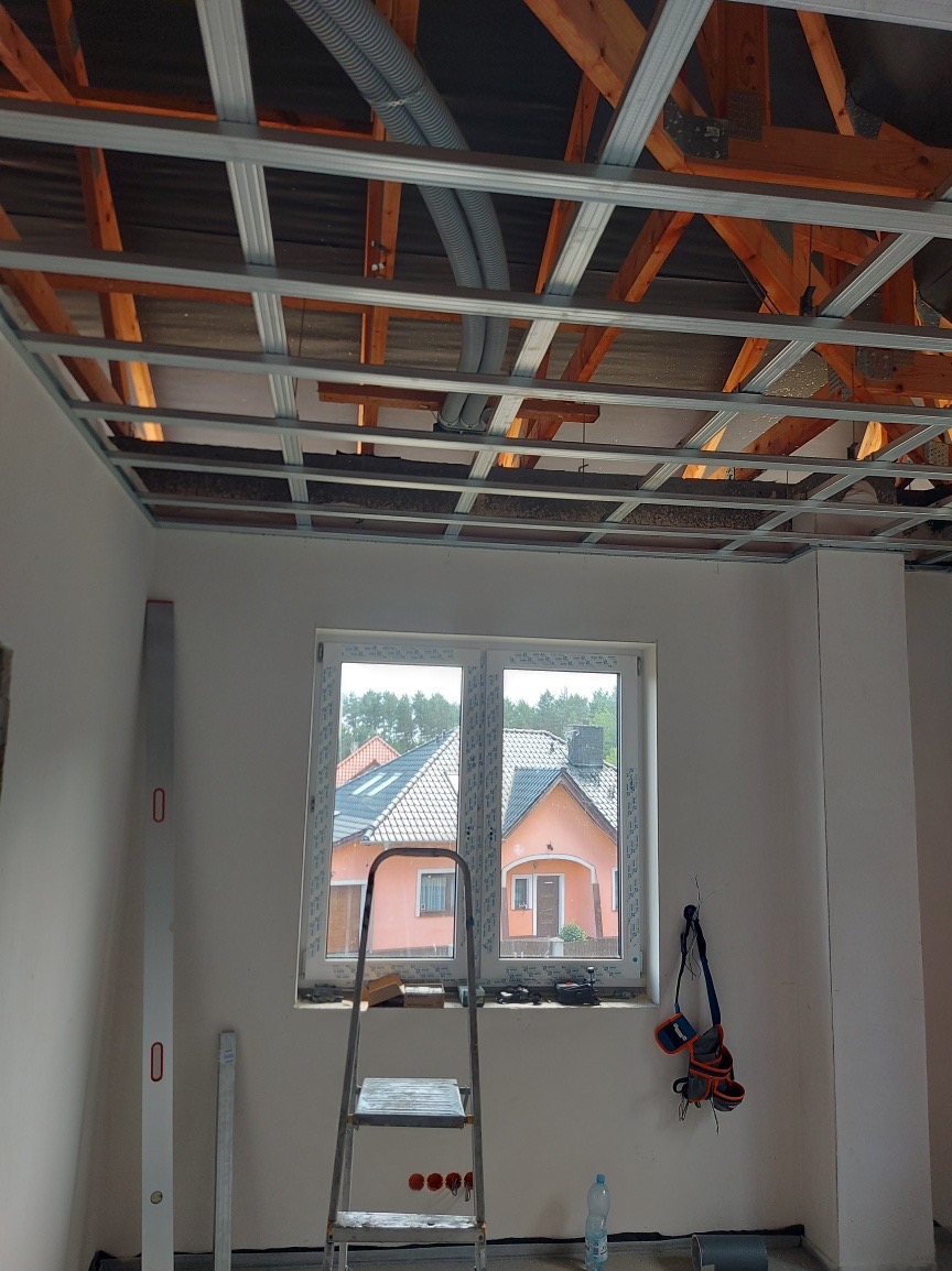 Montaż sufitu podwieszanego z płyt karton-gips: metalowa konstrukcja, widoczne przewody wentylacyjne, drabina, poziomica i okno z widokiem na dom.