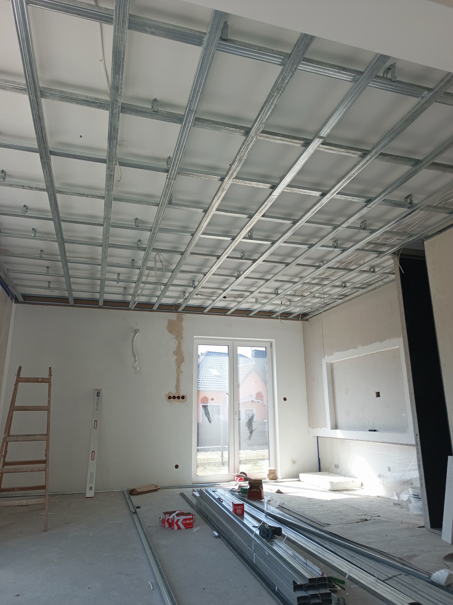 Montaż sufitu podwieszanego z płyt gipsowo-kartonowych. Widoczna metalowa konstrukcja, profile i częściowo zamontowane płyty. W tle okno i ściany w stanie surowym.