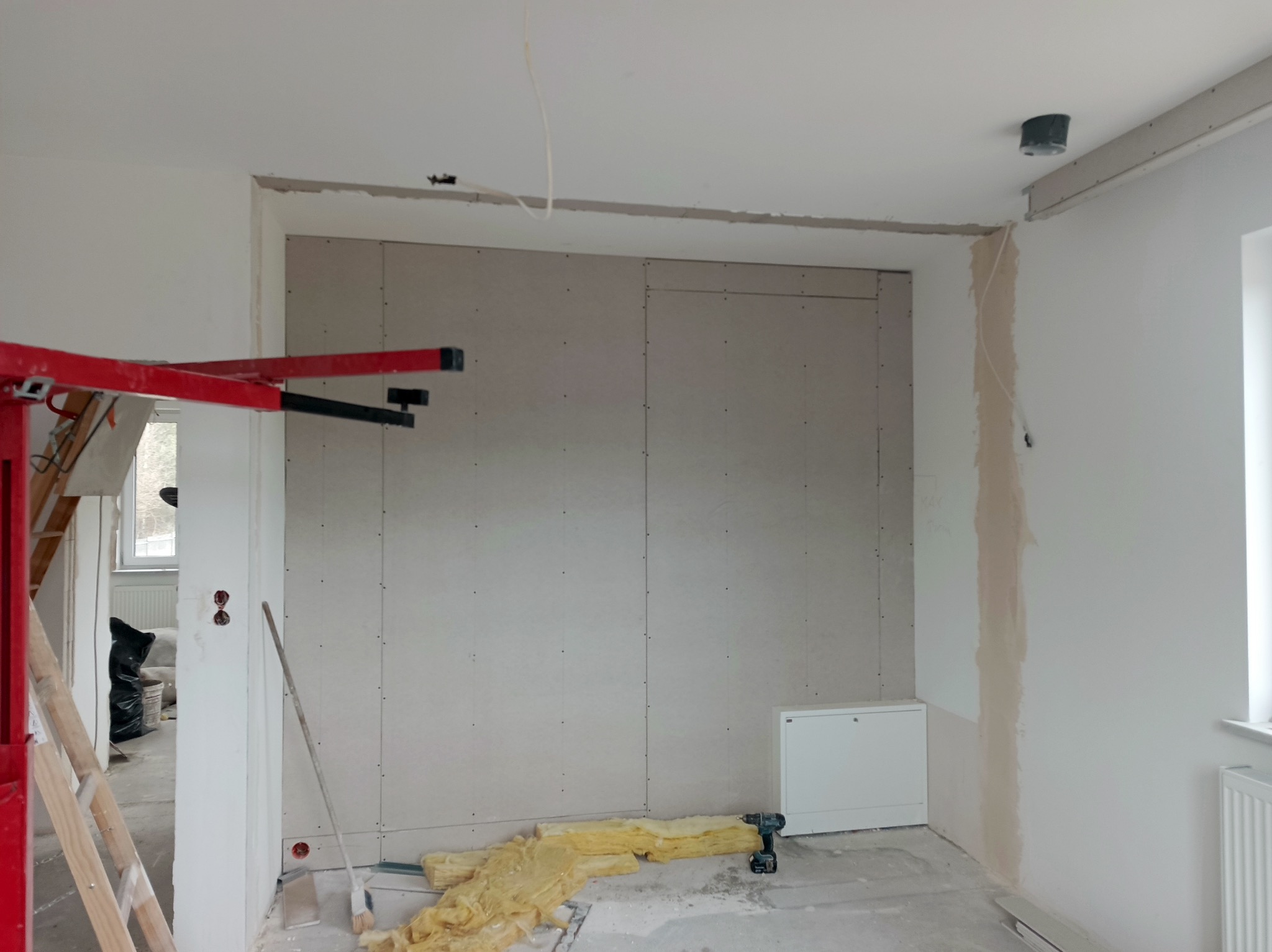 Montaż płyty gipsowo-kartonowej na ścianie z widocznym rusztowaniem, narzędziami i izolacją. Etap prac wykończeniowych w jasnym wnętrzu.