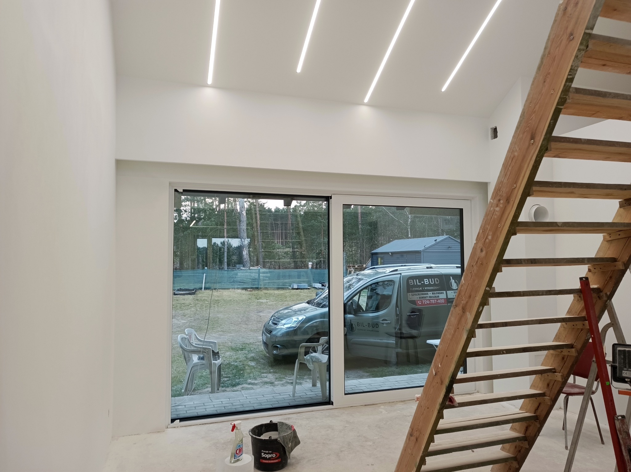 Jasne wnętrze po remoncie: gładkie, białe ściany, nowoczesne oświetlenie liniowe, drewniane schody. Widok na zewnątrz przez duże okno przesuwne.