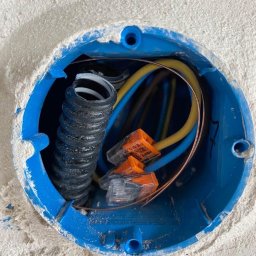 DAWID GASANOW - USŁUGI ELEKTRYCZNE AZPOL - Niebieska puszka instalacyjna w ścianie z przewodami (żółty, niebieski, miedziany) i złączkami WAGO. Widoczna karbowana rurka. Ściana w surowym tynku.